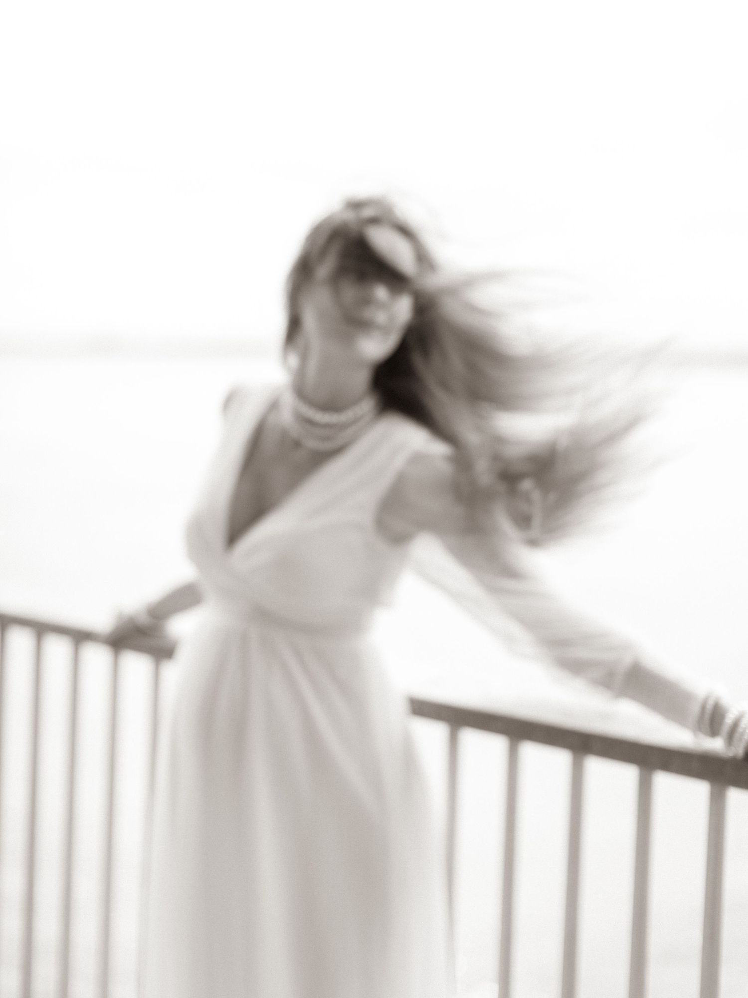 A woman in a white dress stands on a railing, her hair blowing in the wind in philly, in a dreamy black and white photo.