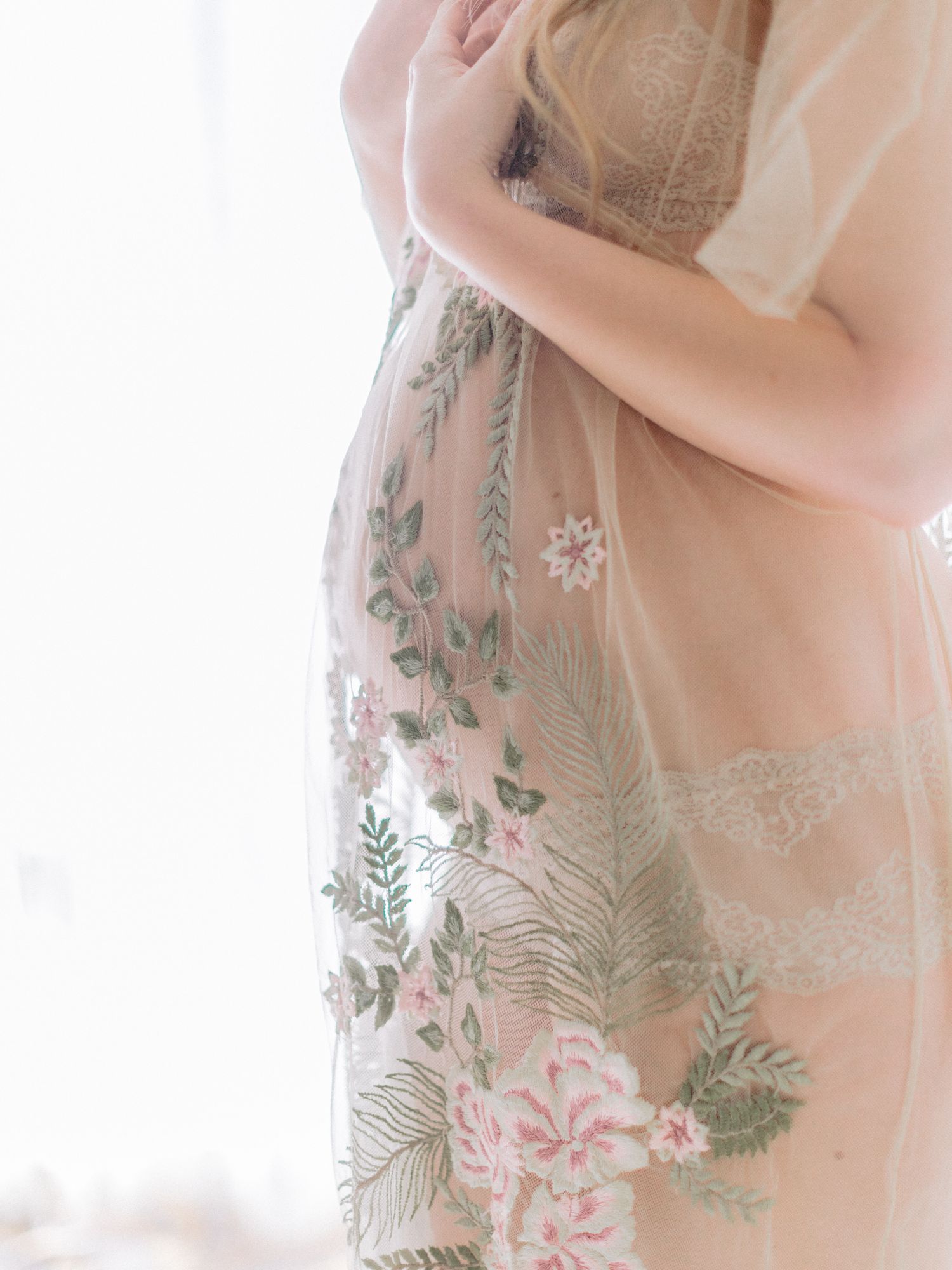 Pregnant woman in sheer floral embroidered maternity gown with pink roses and green leaves near bright window.