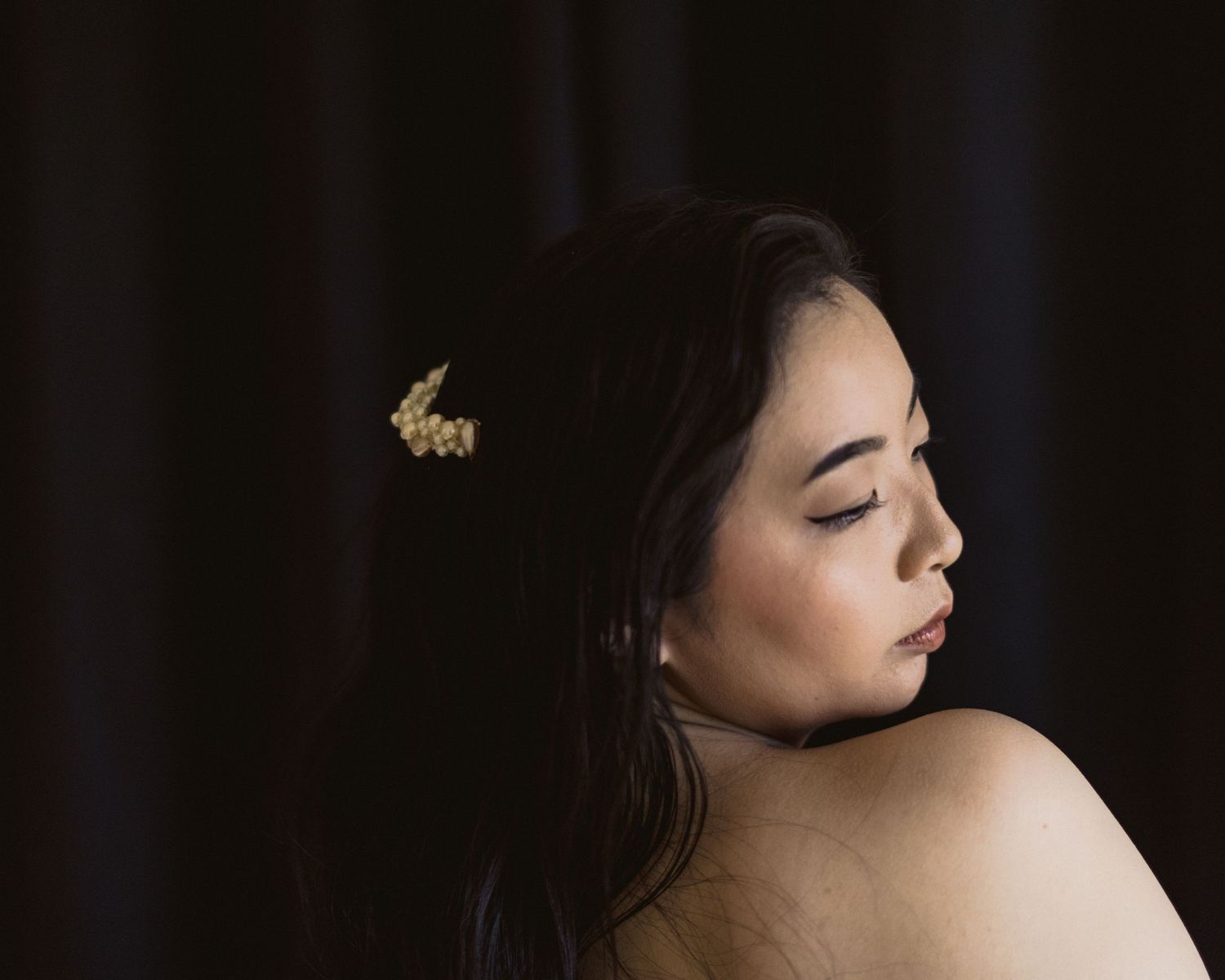 Young woman in profile with floral hair accessory against dark background, bare shoulder, elegant portrait photography.