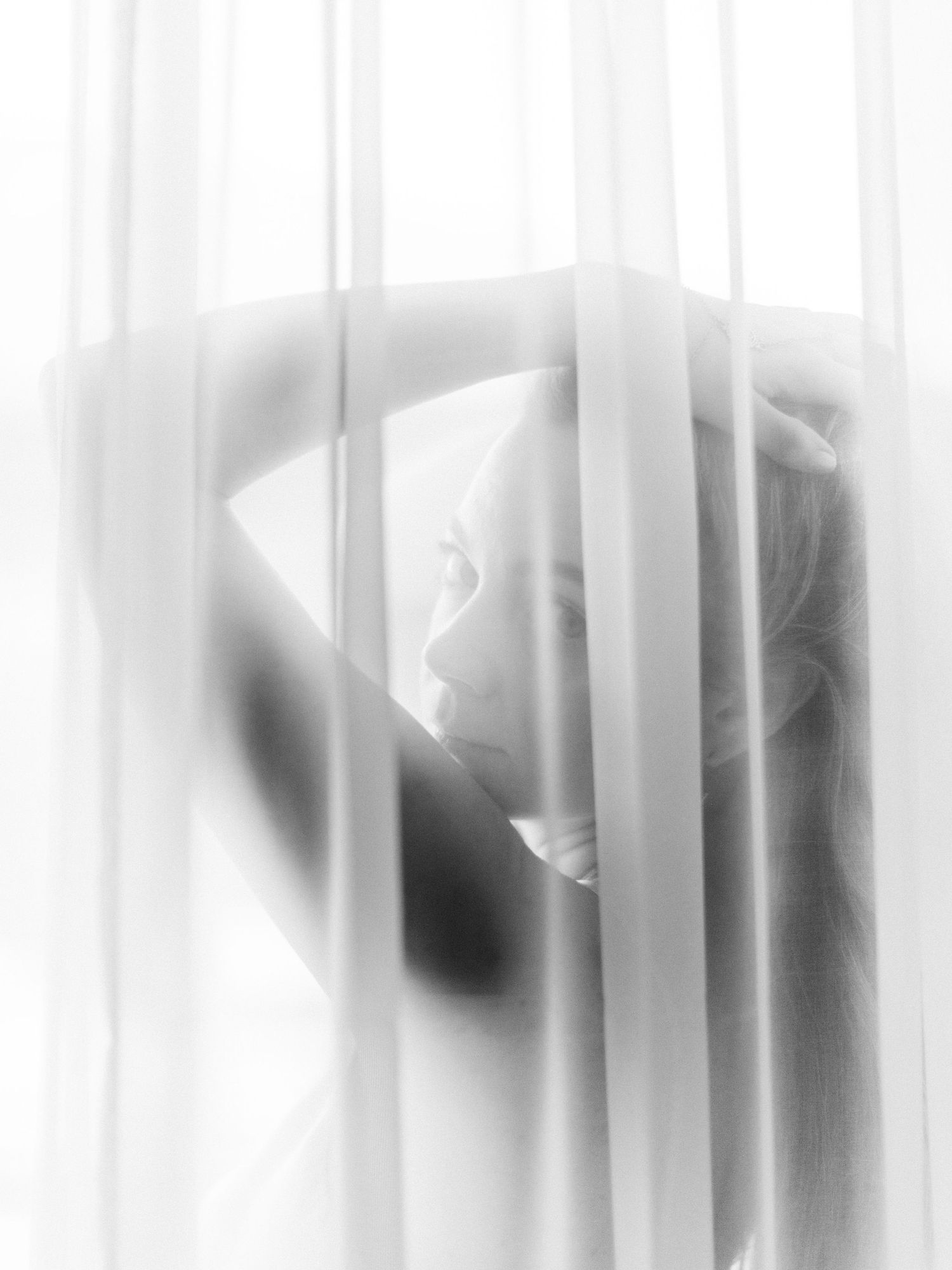 Black and white artistic photo of a woman silhouetted behind sheer white curtains in soft diffused light.