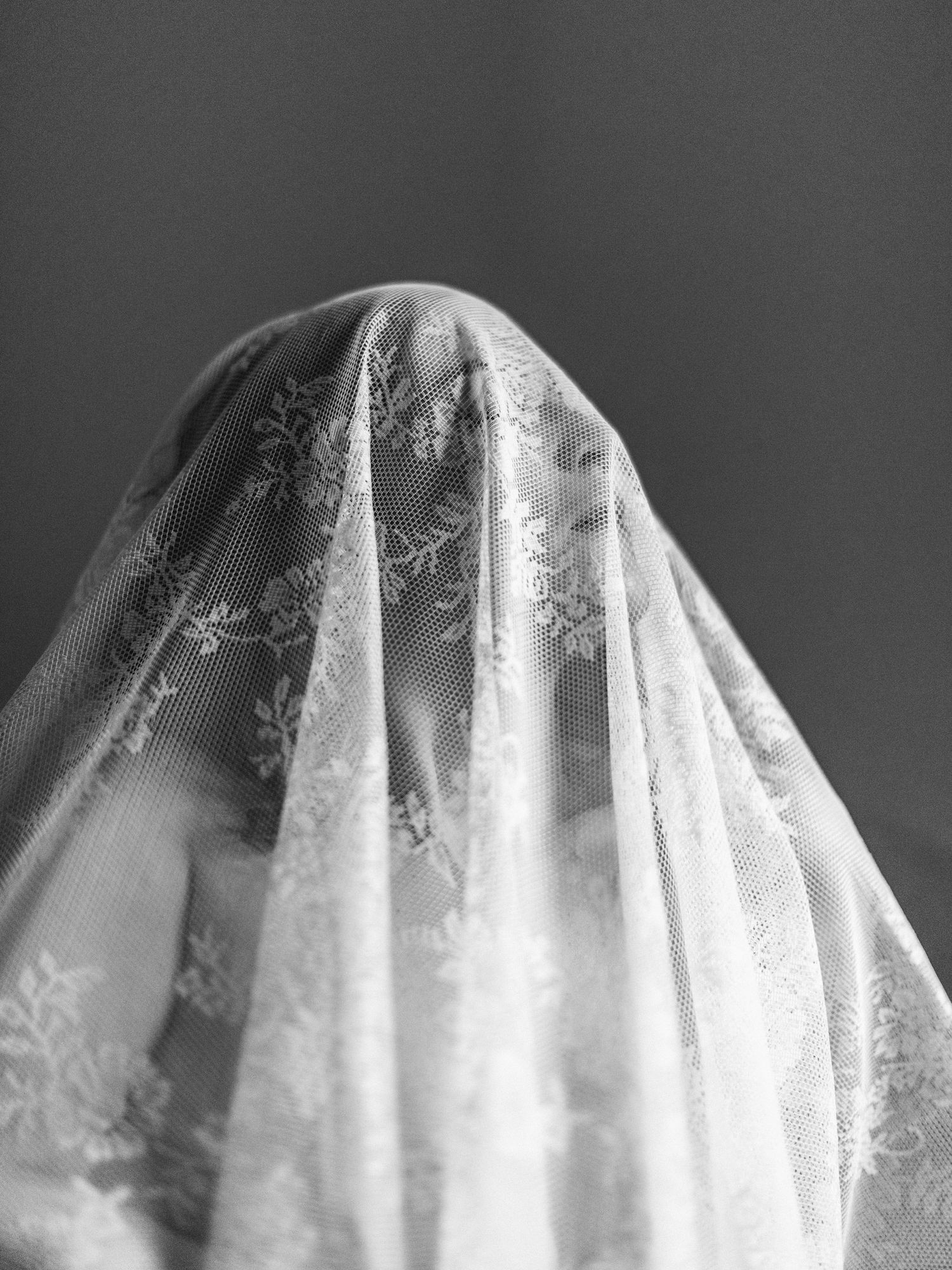 Black and white photo of a person draped in delicate floral lace wedding veil, obscuring their face.
