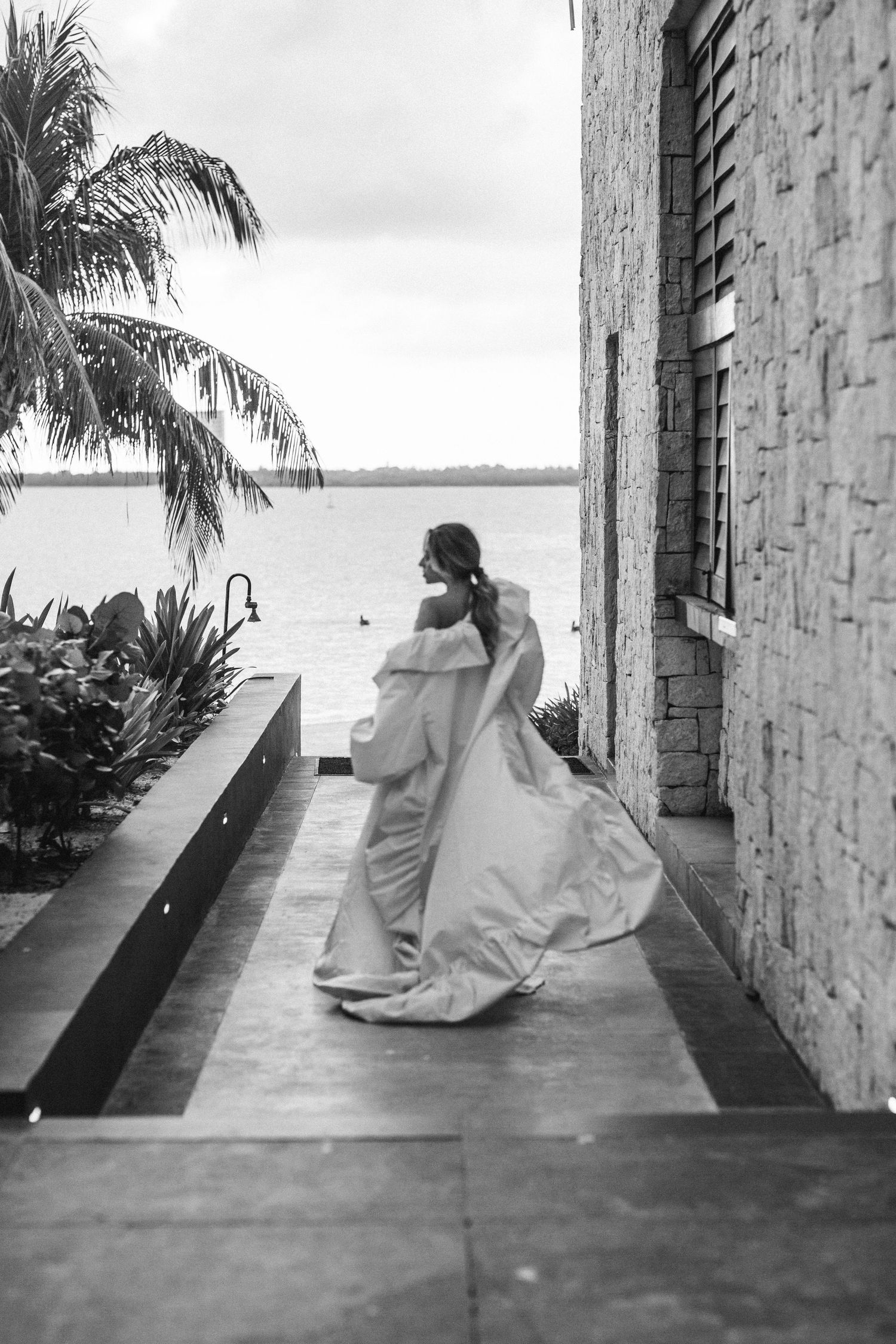 Bride in elegant white gown walking along waterfront path with palm trees in black and white photo.
