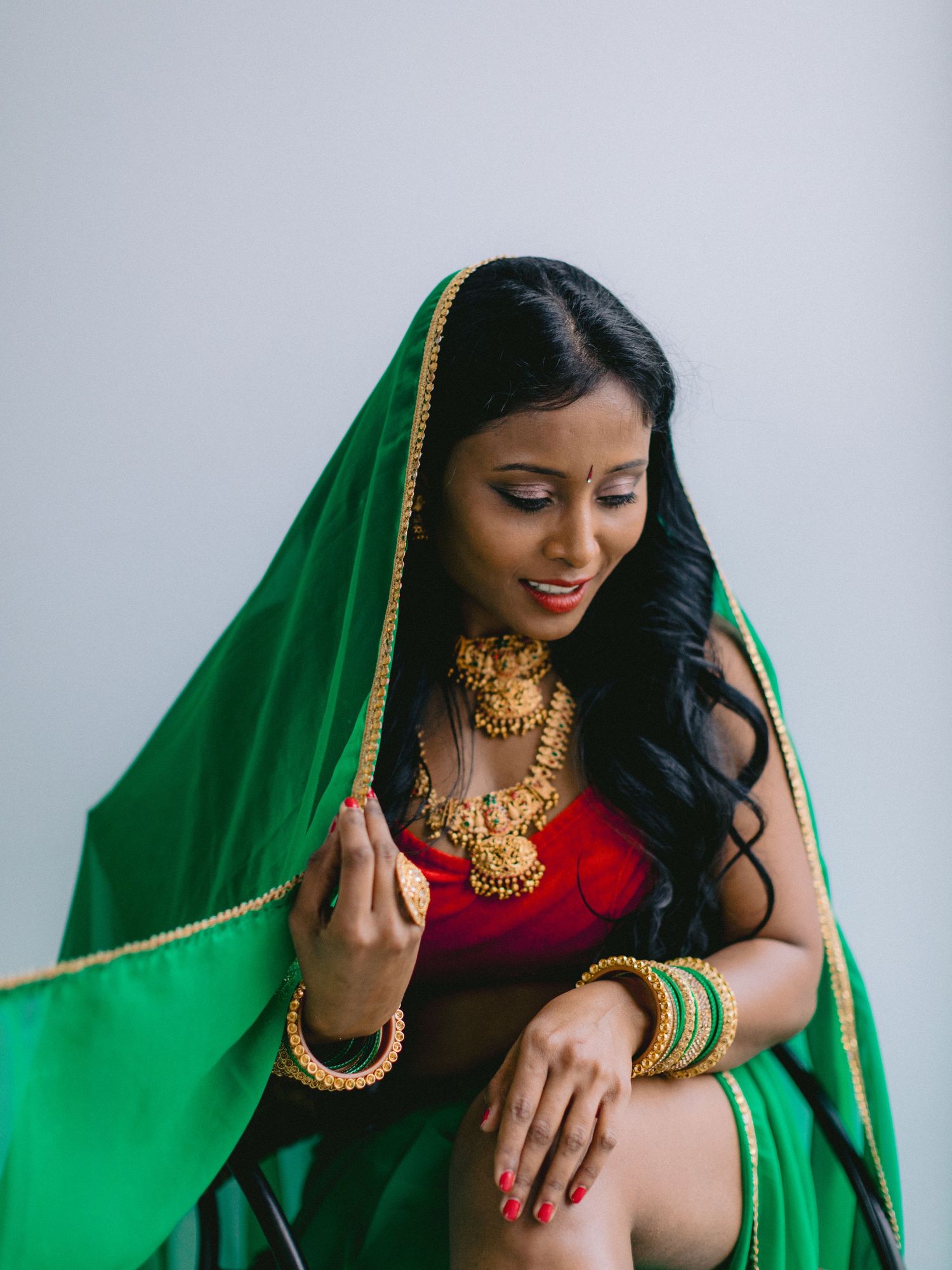 Woman in traditional South Asian attire wearing red and green sari with gold jewelry poses elegantly against gray background.