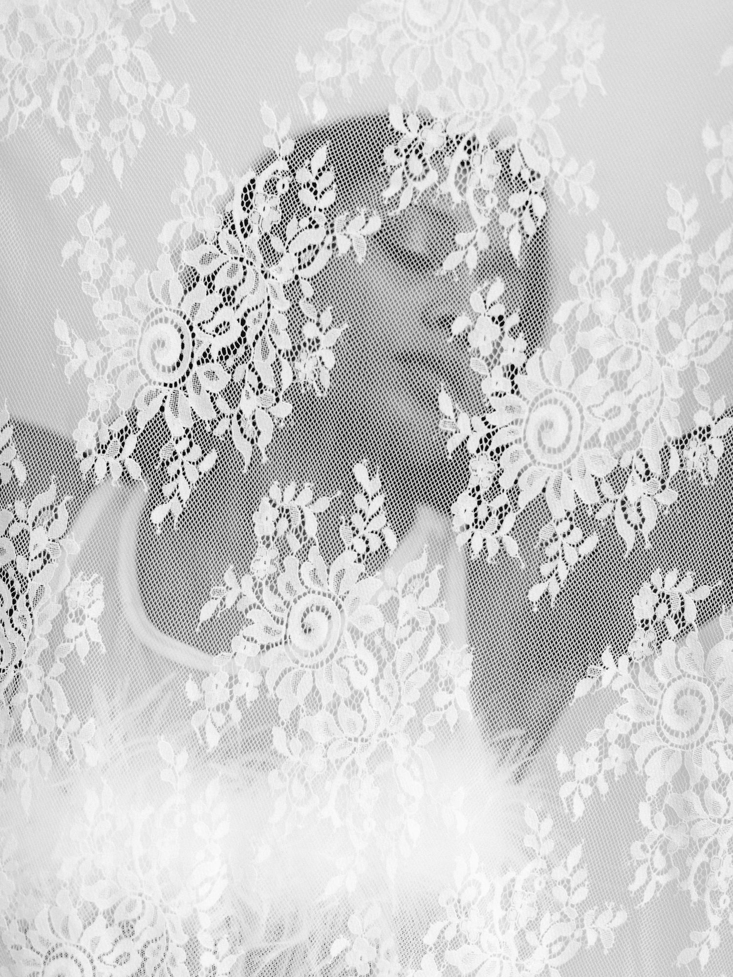 A black and white close-up photograph through delicate white lace fabric creating an ethereal, romantic pattern effect.