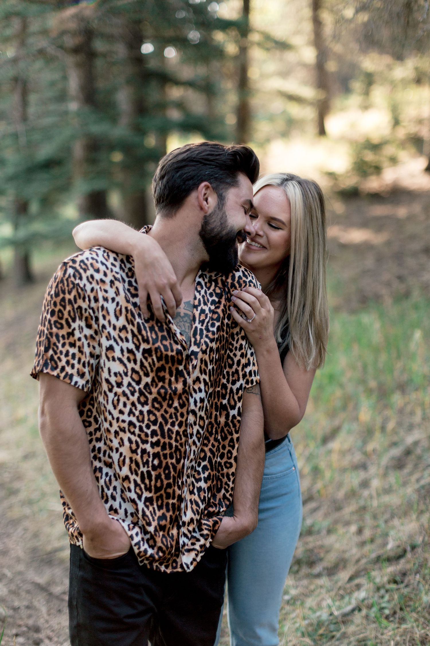 Smiling blonde woman embracing bearded man in leopard print shirt in a forest setting.