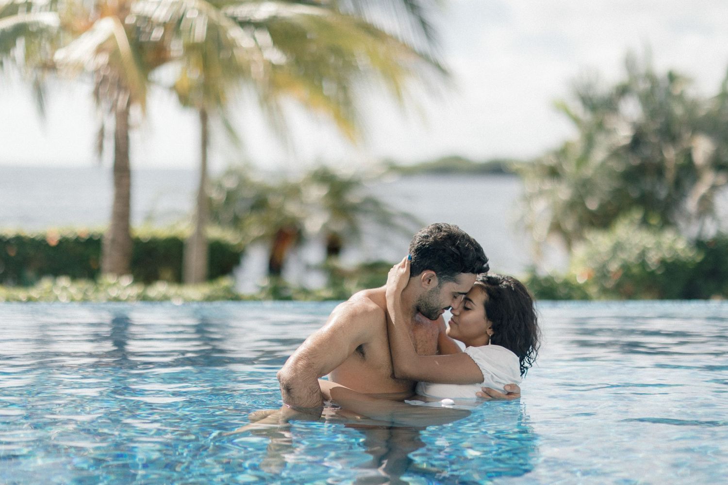 Romantic couple embracing and kissing in tropical infinity pool surrounded by palm trees.