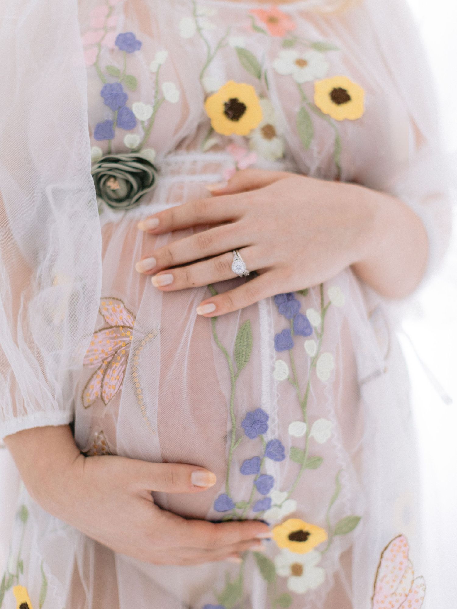 Close-up of pregnant belly in floral embroidered sheer dress with diamond engagement ring.
