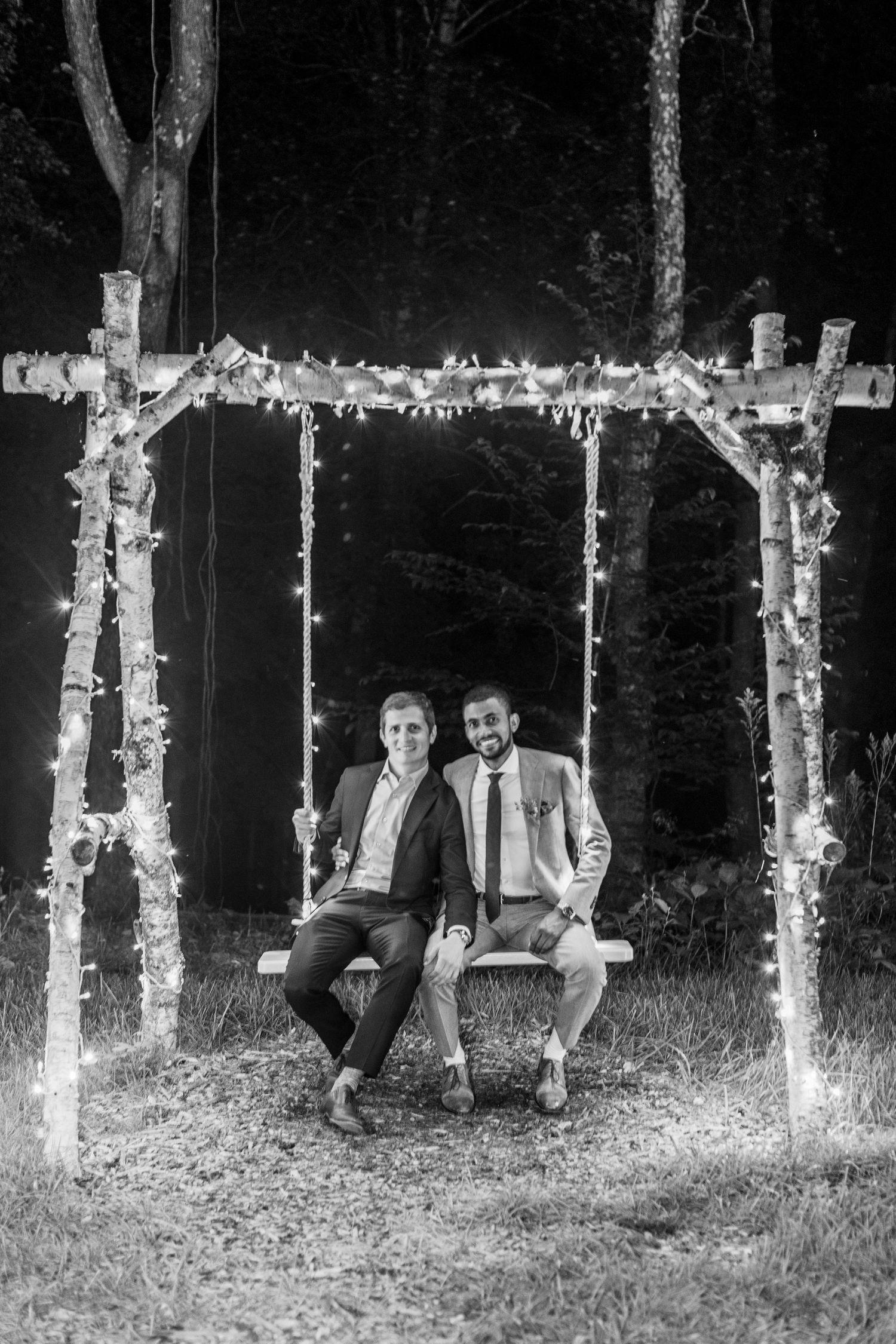 Black and white photo of two grooms sitting on a swing adorned with fairy lights.