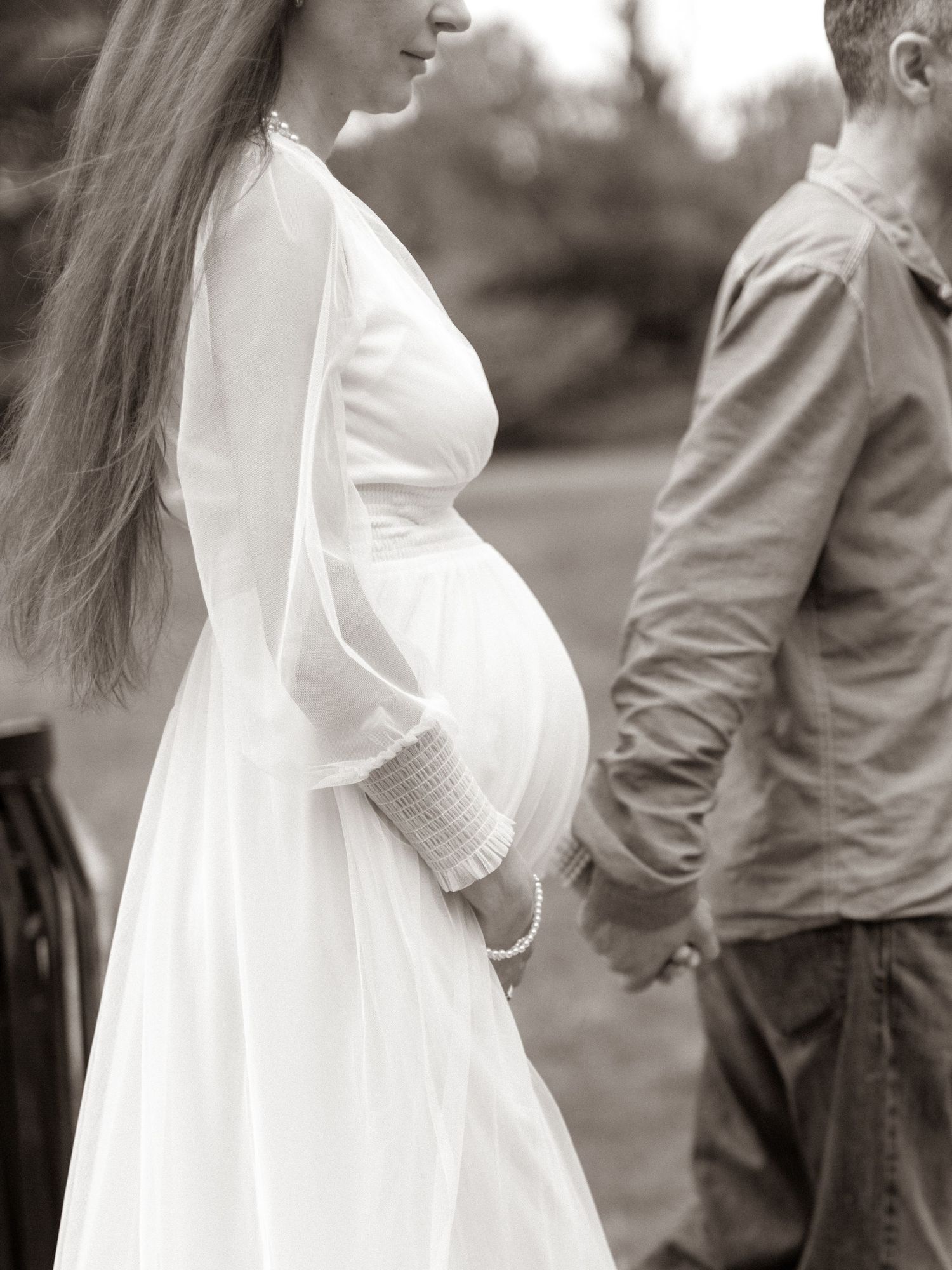 Black and white photo of pregnant woman in white dress holding hands with partner outdoors.