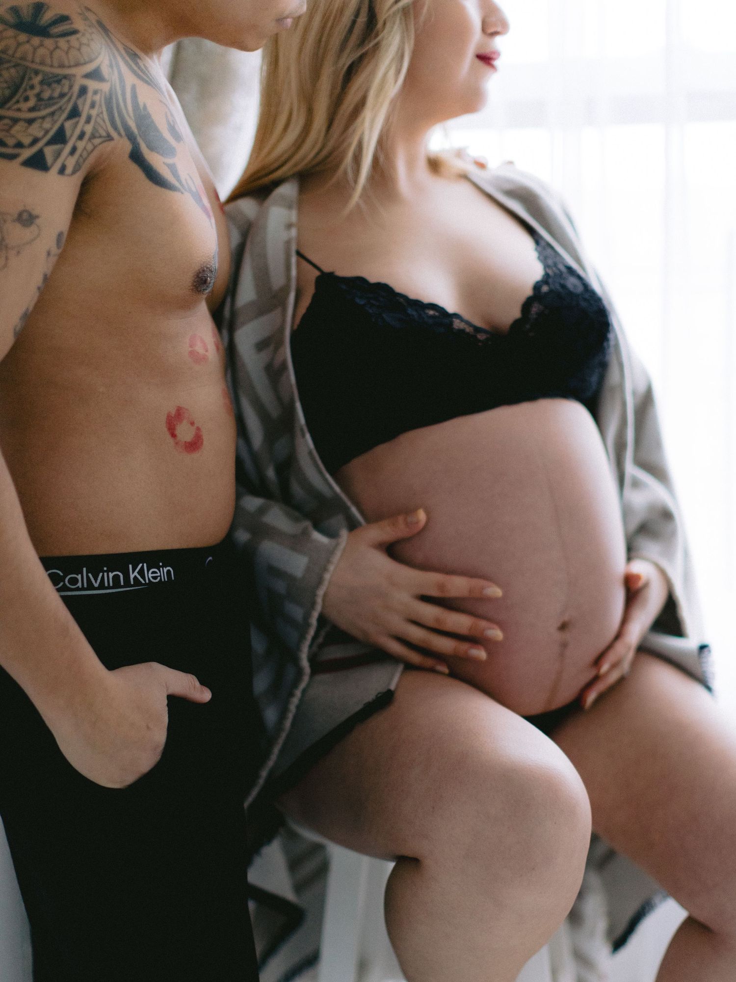Pregnant woman with partner posing intimately, man in Calvin Klein underwear with lipstick mark, woman in black lace bra cradling baby bump.