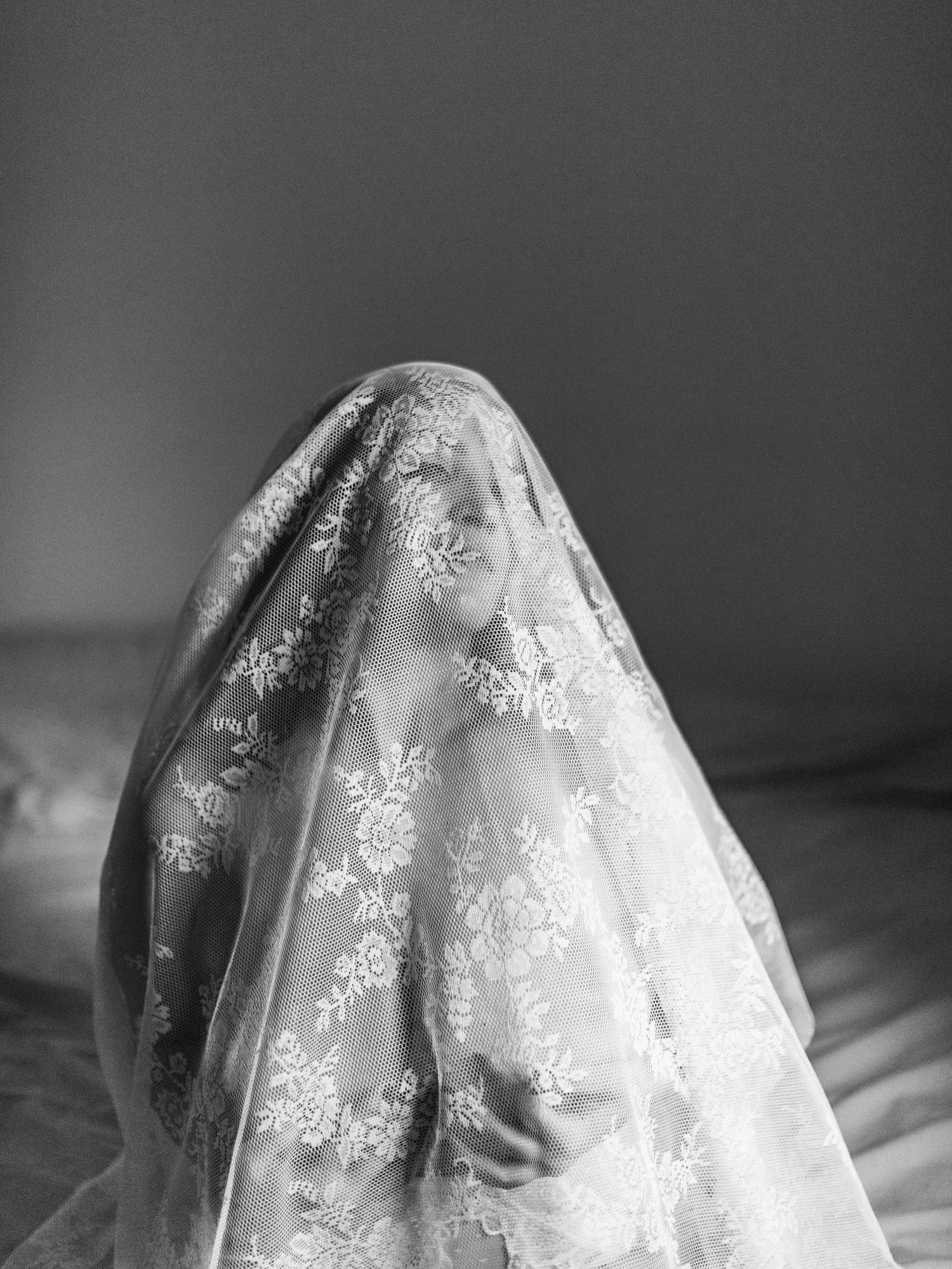 A black and white photograph shows a delicate lace veil with floral patterns draped across a woman.
