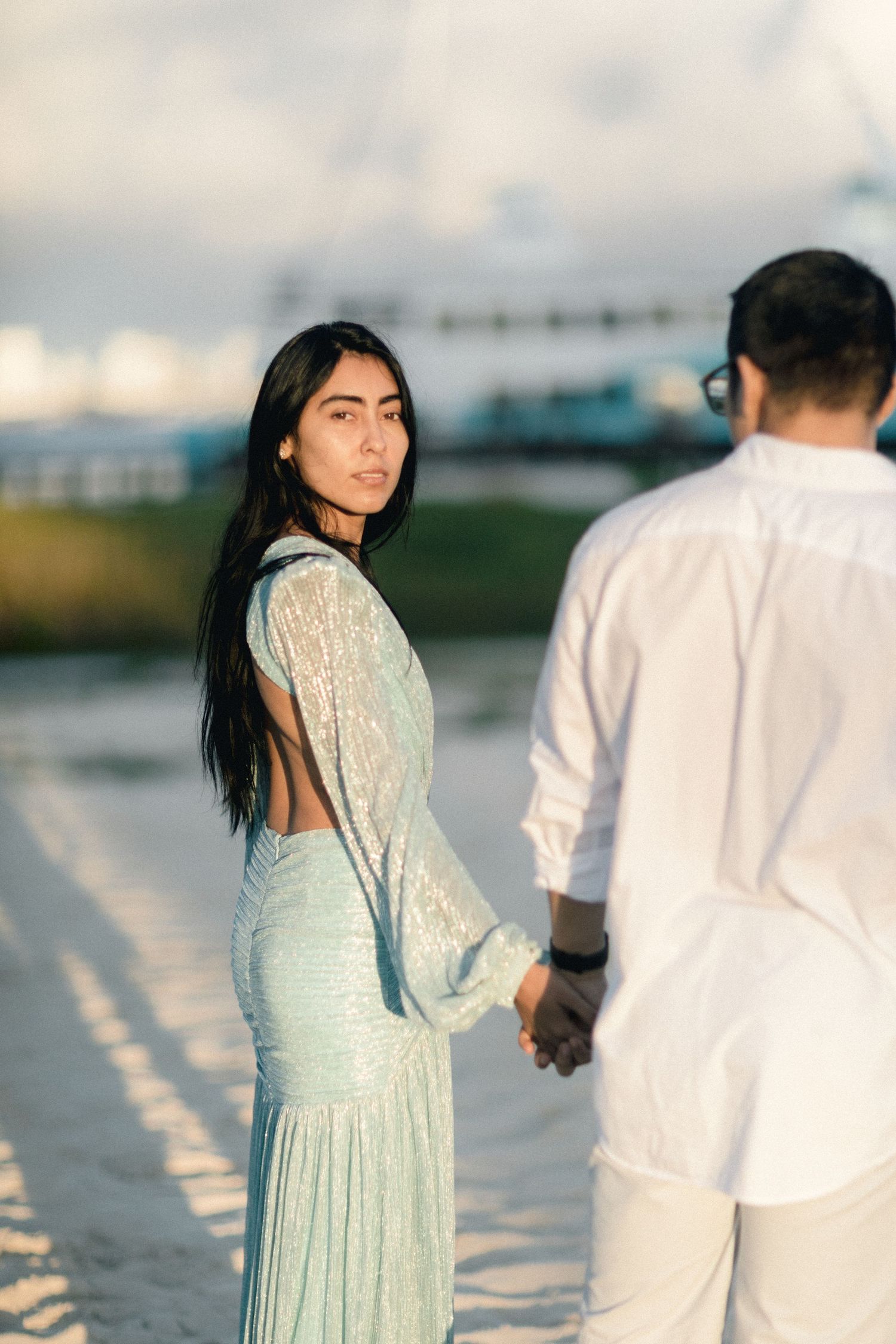 Elegant woman in backless mint sequin gown holds hands with man while looking back on beach.