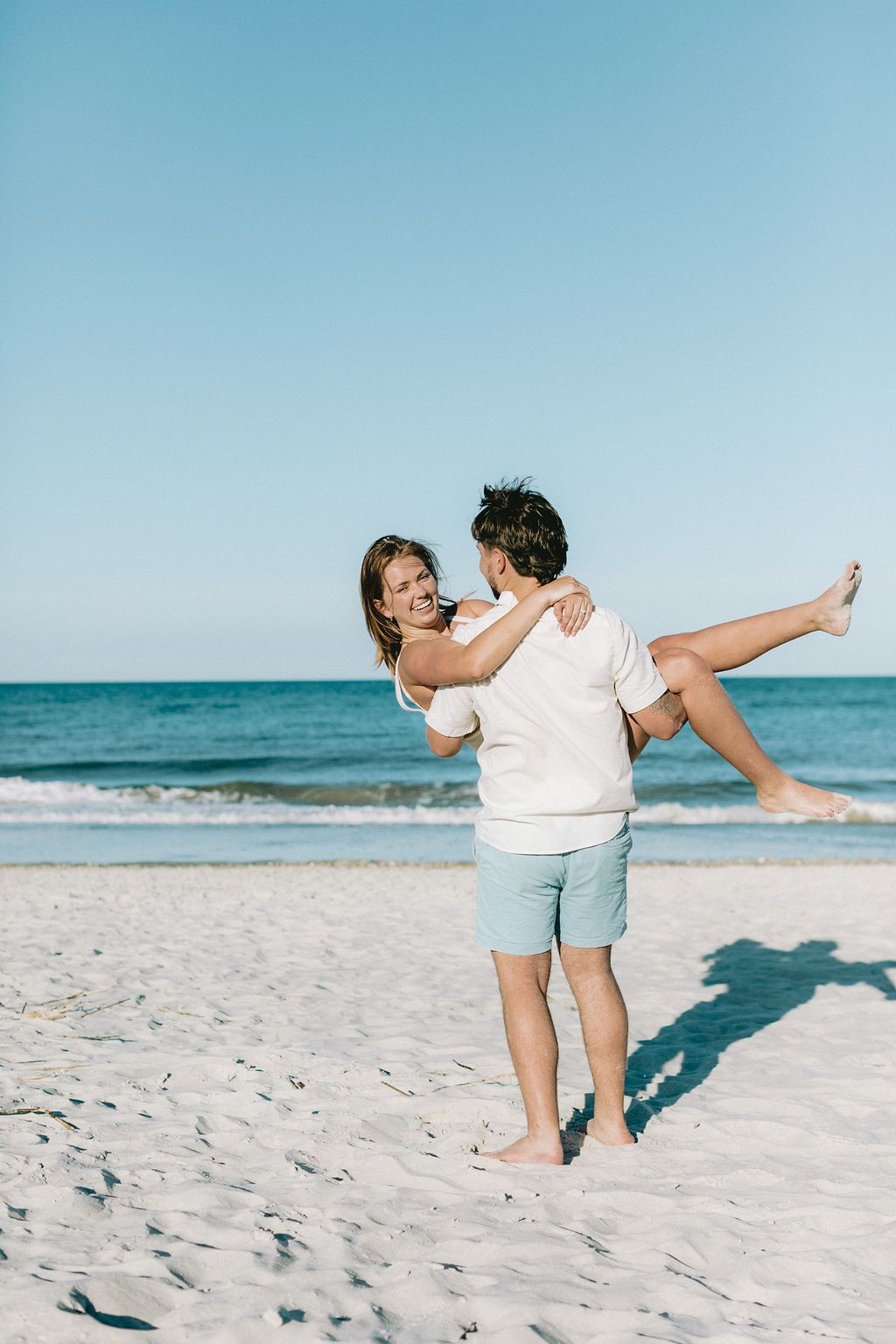 Man joyfully lifting smiling woman on white sandy beach with clear blue ocean in background.