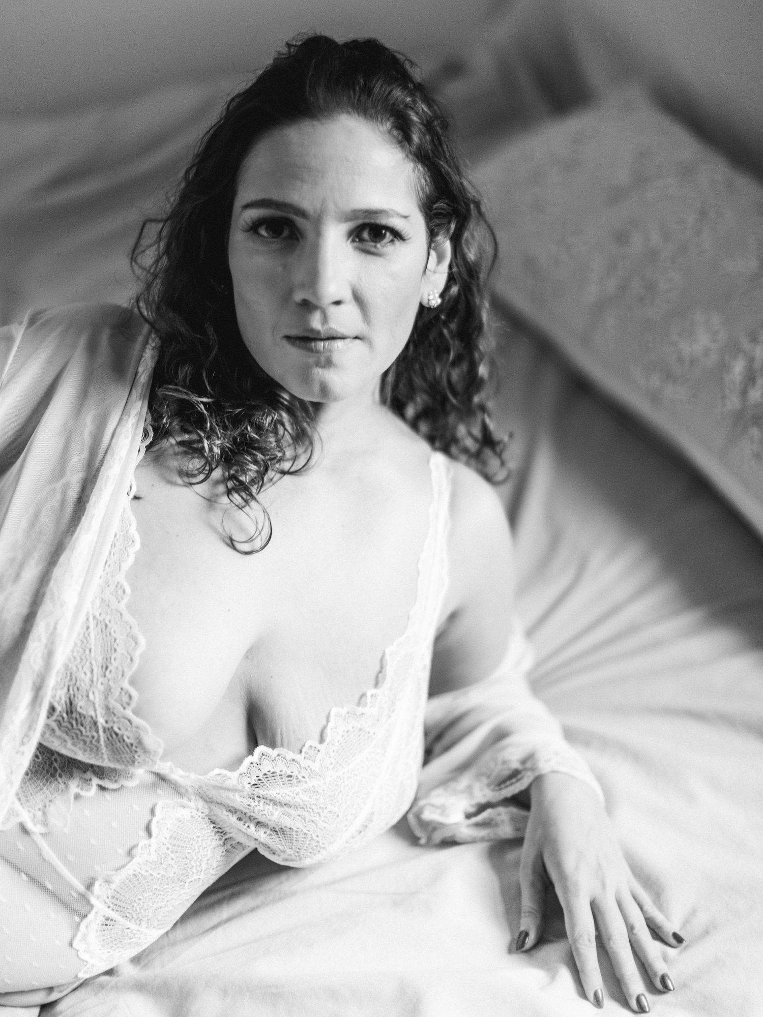 A black and white boudoir photo of a woman showing intimate poses in white lace lingerie against light bedding.