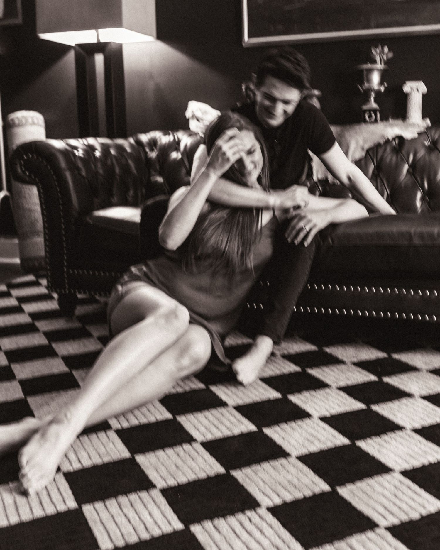 Black and white photo of couple playing on checkered floor near leather sofa indoors.