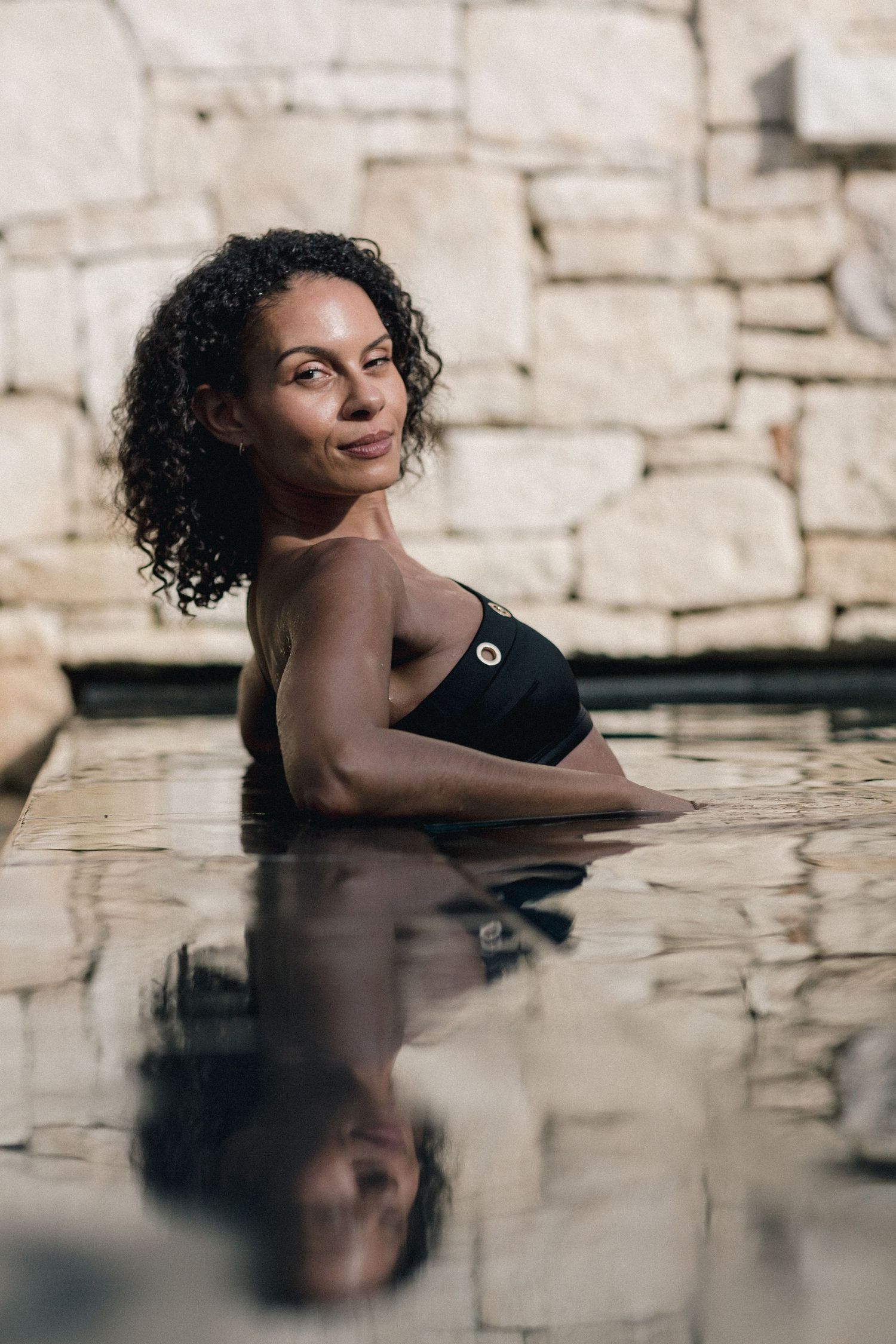 Woman in black swimsuit relaxing in a pool with stone wall backdrop and serene water reflection.
