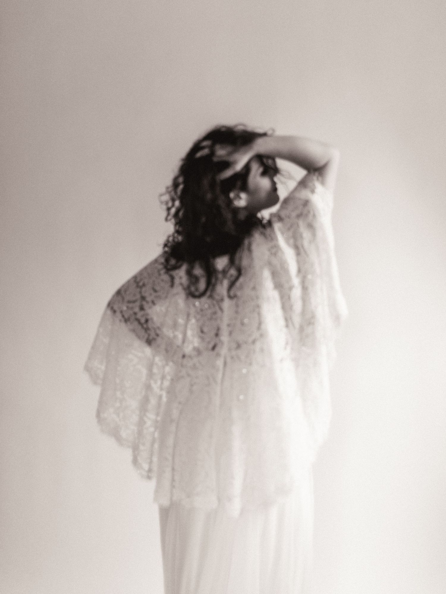Woman in white lace bridal cape and gown poses elegantly in black and white portrait photography.