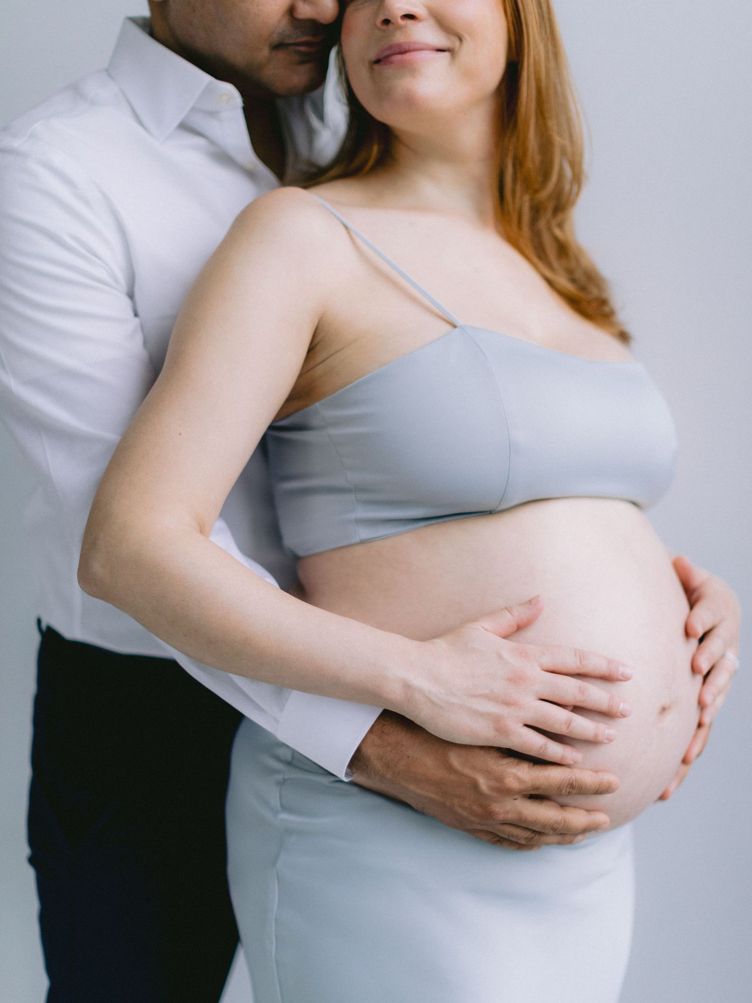 Couple tenderly cradling pregnant belly in a studio, woman in light blue maternity crop top smiling softly.