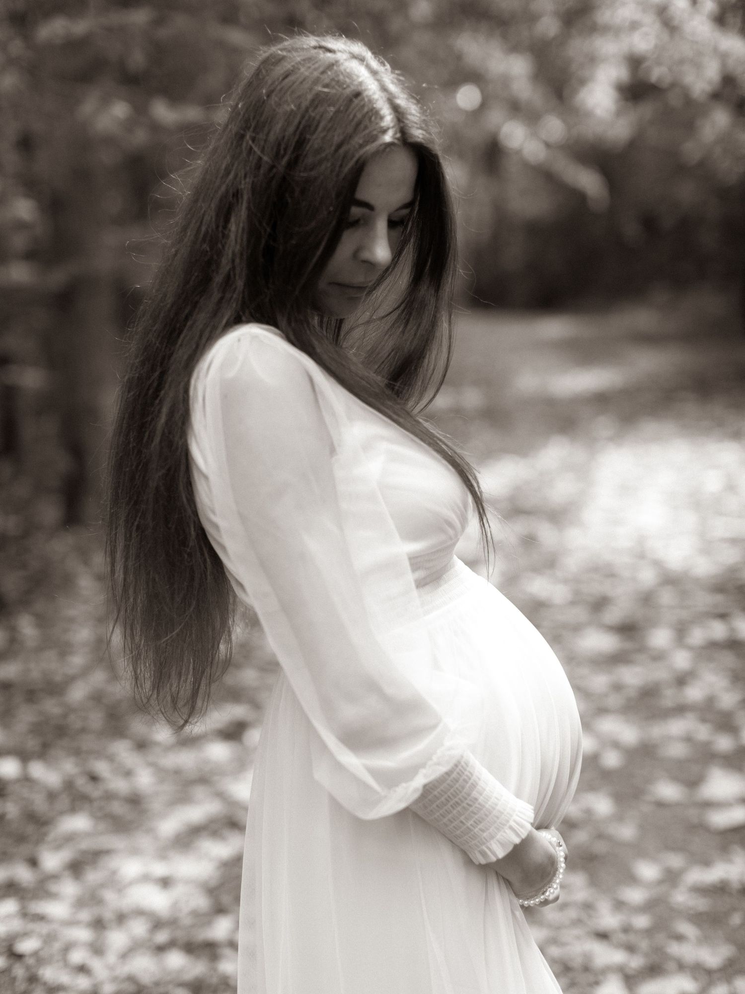 Pregnant woman in white dress cradles baby bump while standing in autumn forest path in sepia maternity portrait.