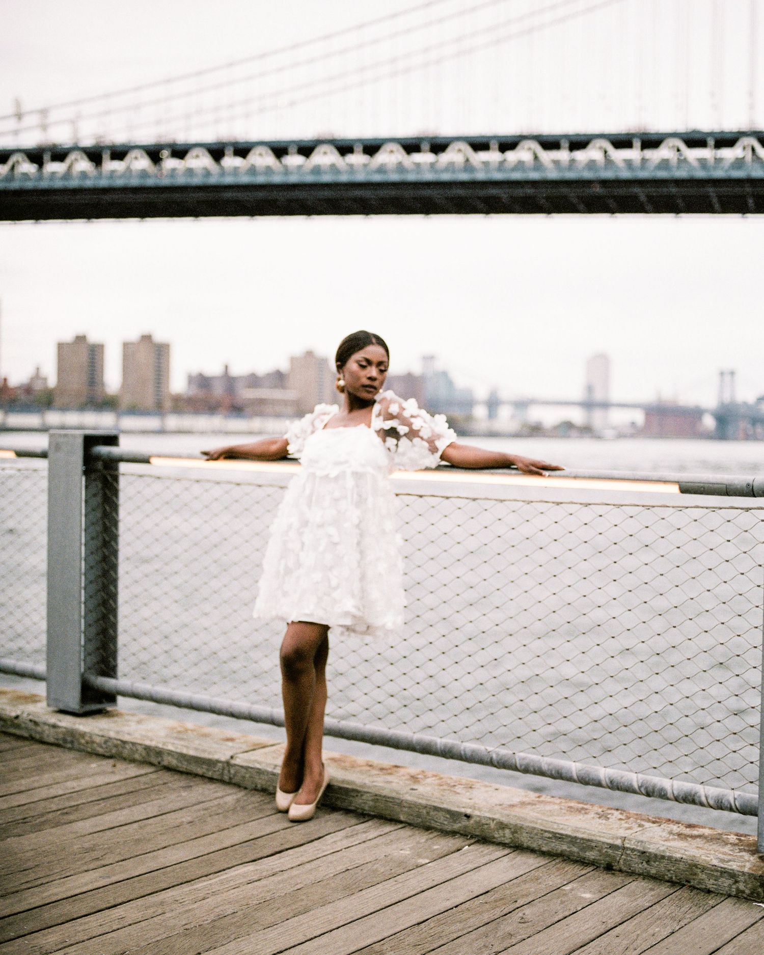 Woman in white mini dress posing on waterfront pier with Manhattan Bridge in background.