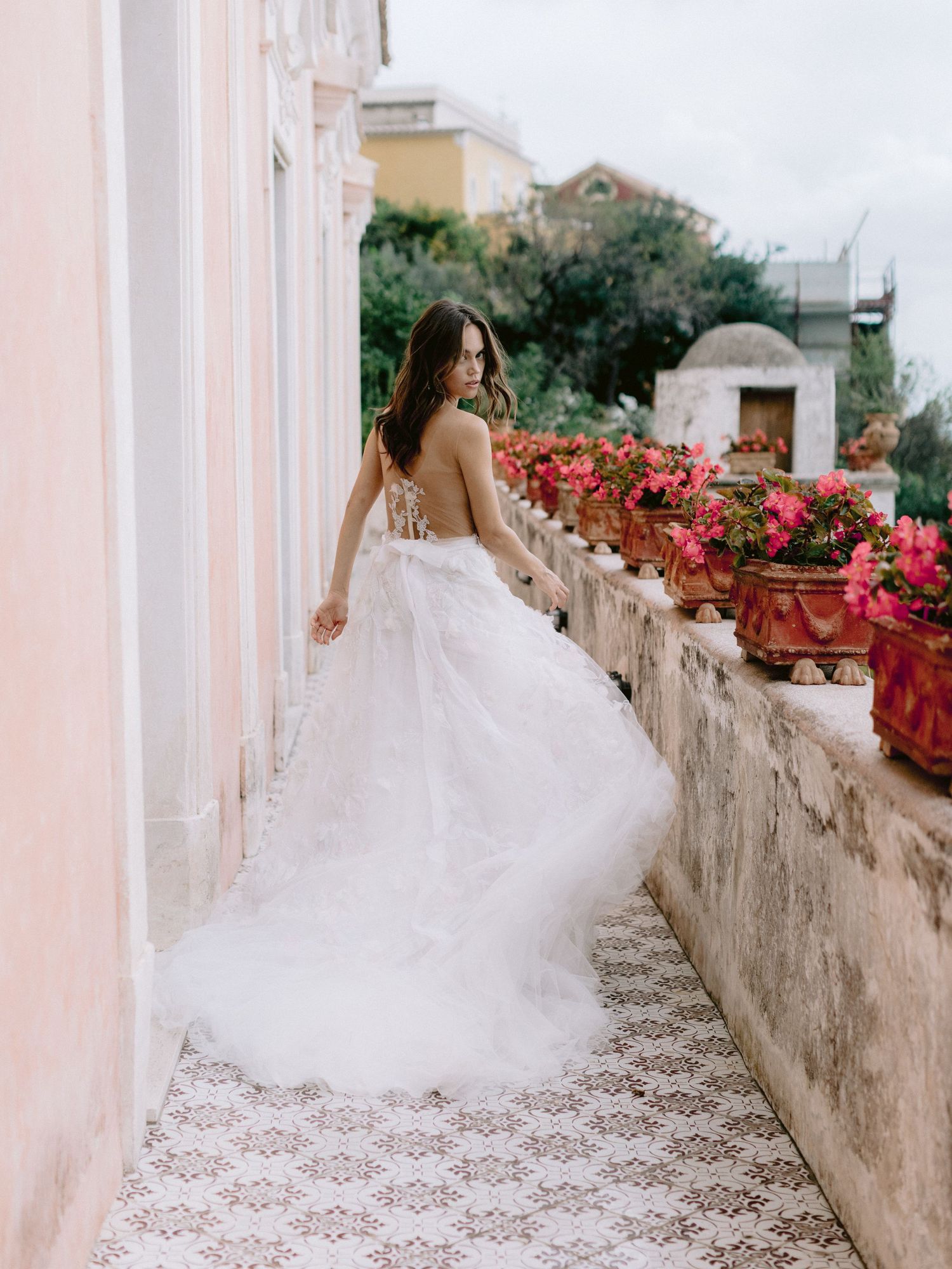 Bride in a flowing white tulle gown on a colorful Mediterranean terrace with pink flowers.