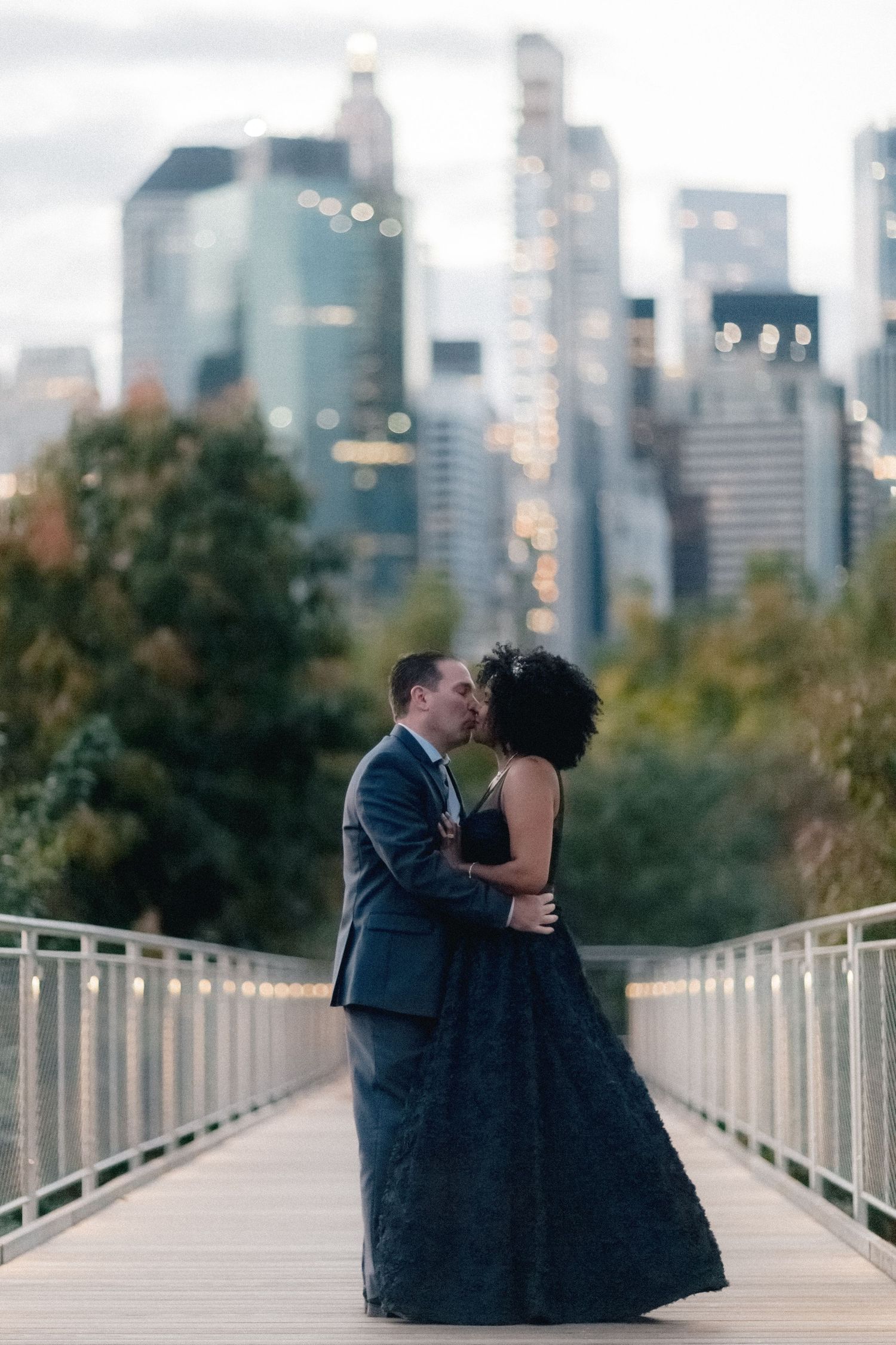 Couple sharing romantic kiss on bridge with illuminated New York City skyline at dusk.