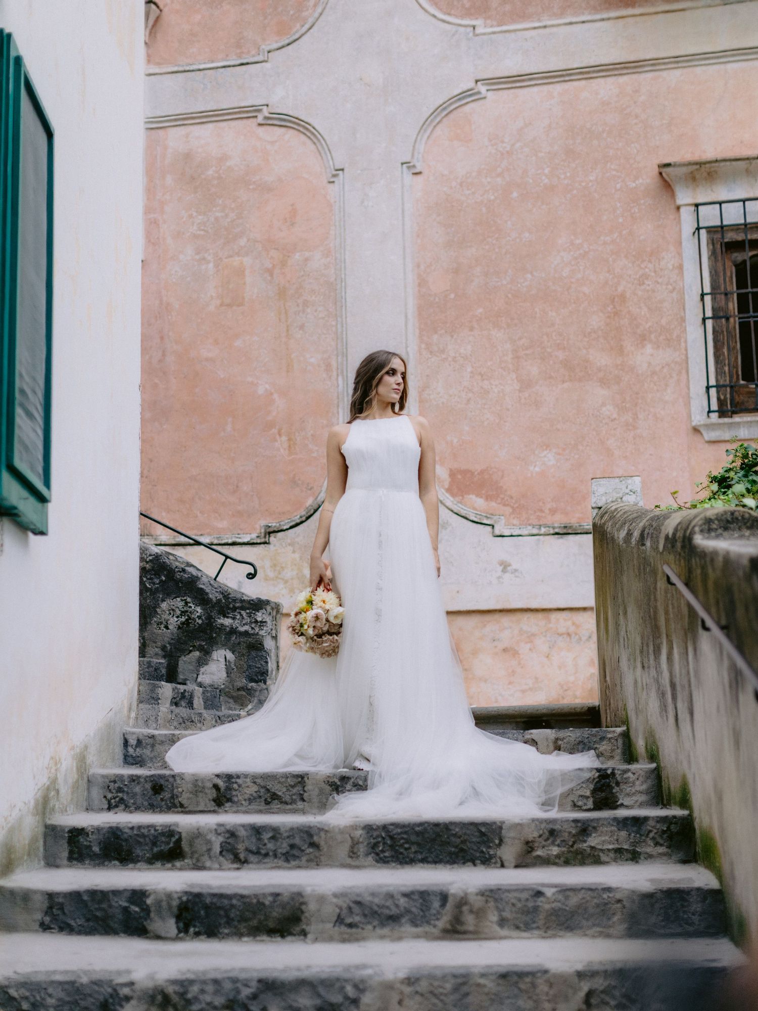 Bride in elegant white gown holding bouquet standing on stone steps of pink Italian building.