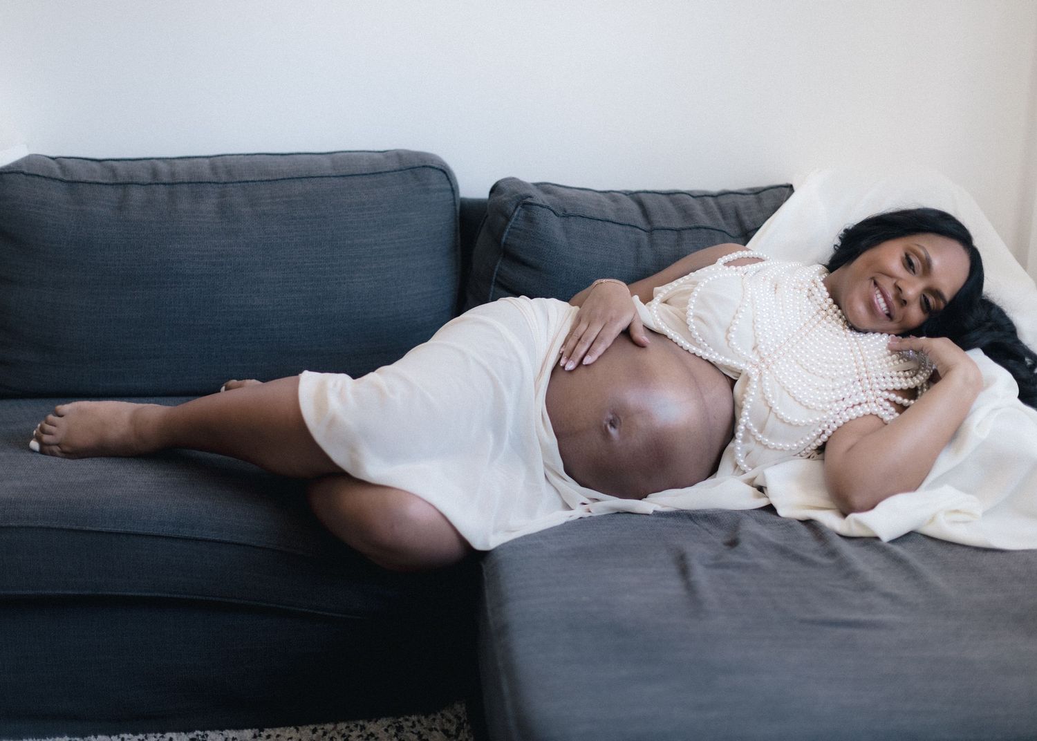 Pregnant woman in white dress resting on gray couch, cradling her baby bump while lying on her side.