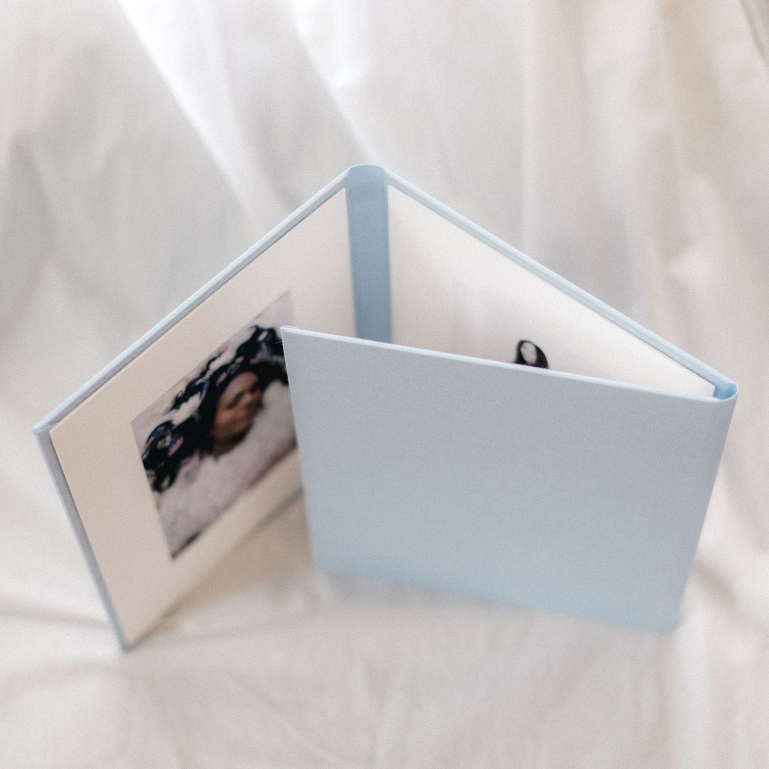 Light blue hardcover photo album open on white fabric, displaying a newborn baby portrait on the left page.