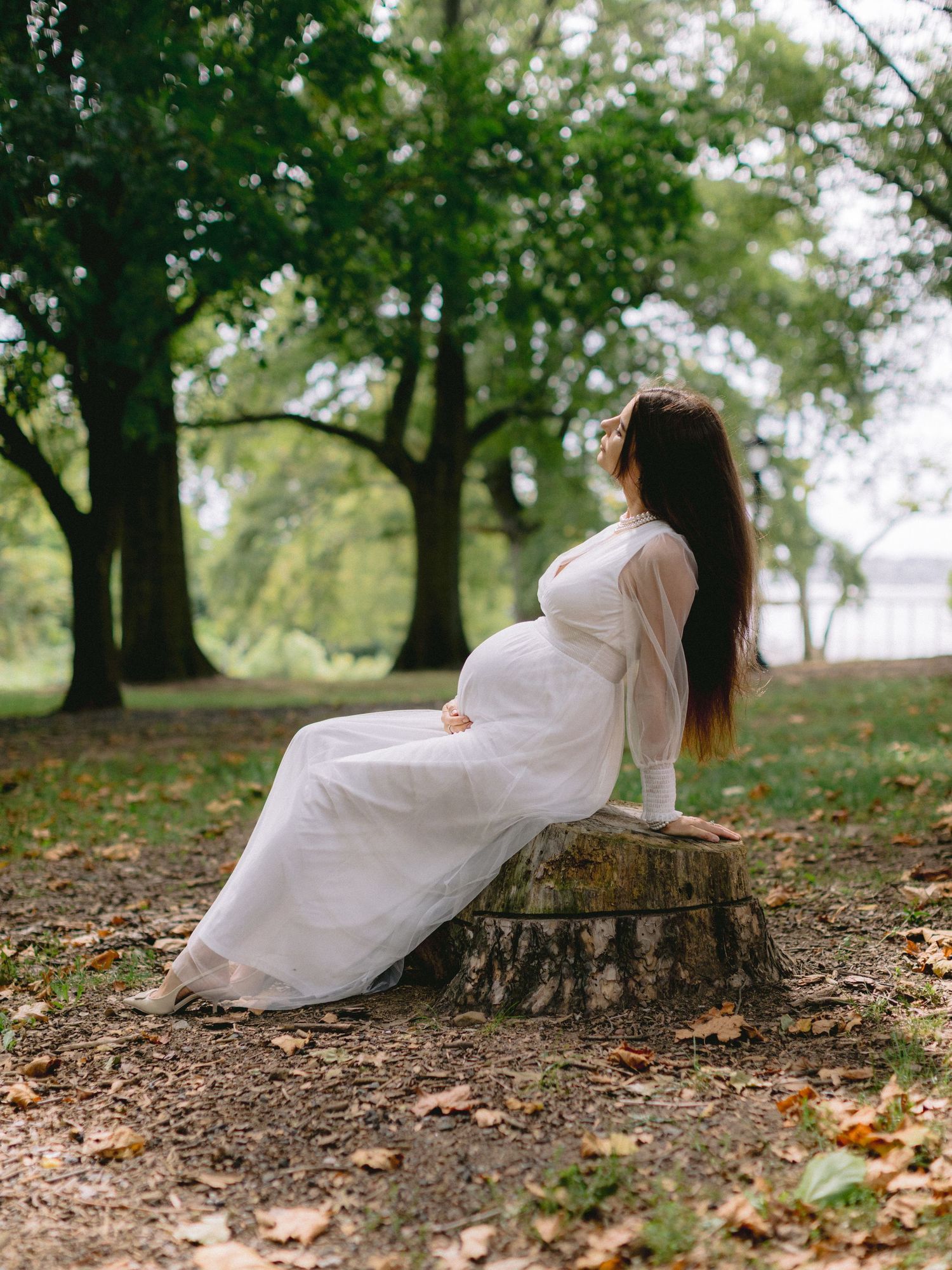 Pregnant woman in white dress sitting on tree stump in lush green park, side profile with long dark hair.