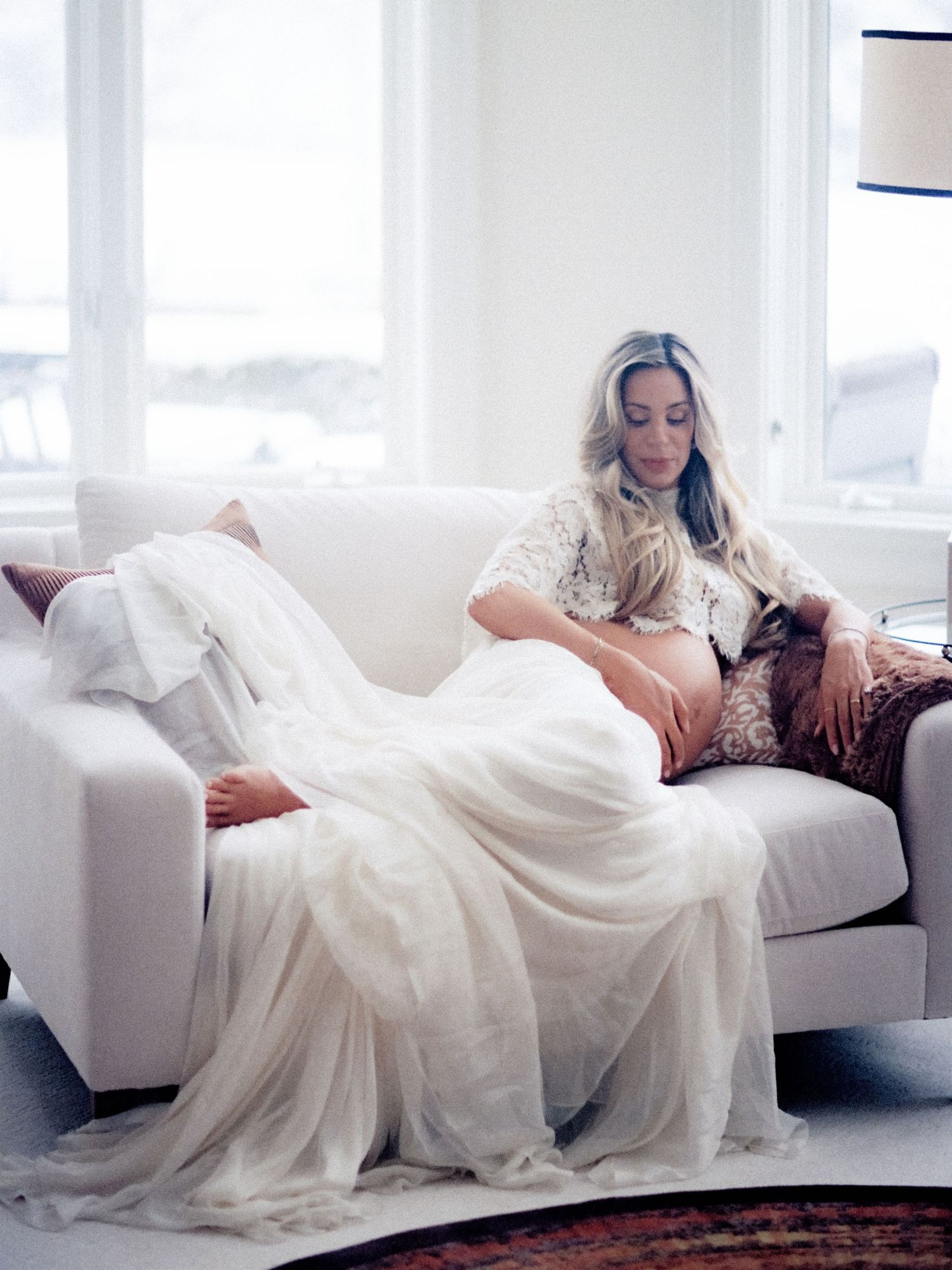 Pregnant woman in white lace top resting on a white sofa, draped in sheer fabric, gazing down at her baby bump.