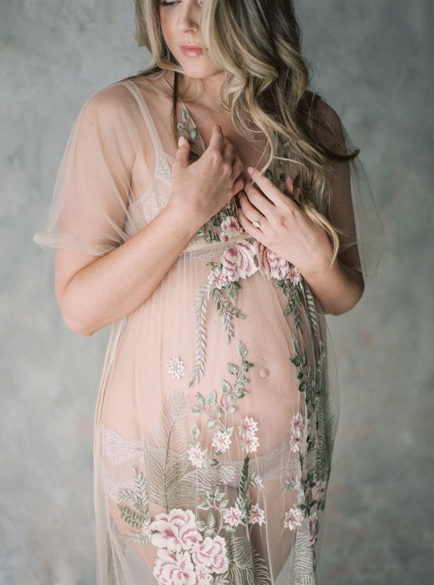 Woman in sheer floral embroidered blush robe posing gracefully against a soft gray studio background.