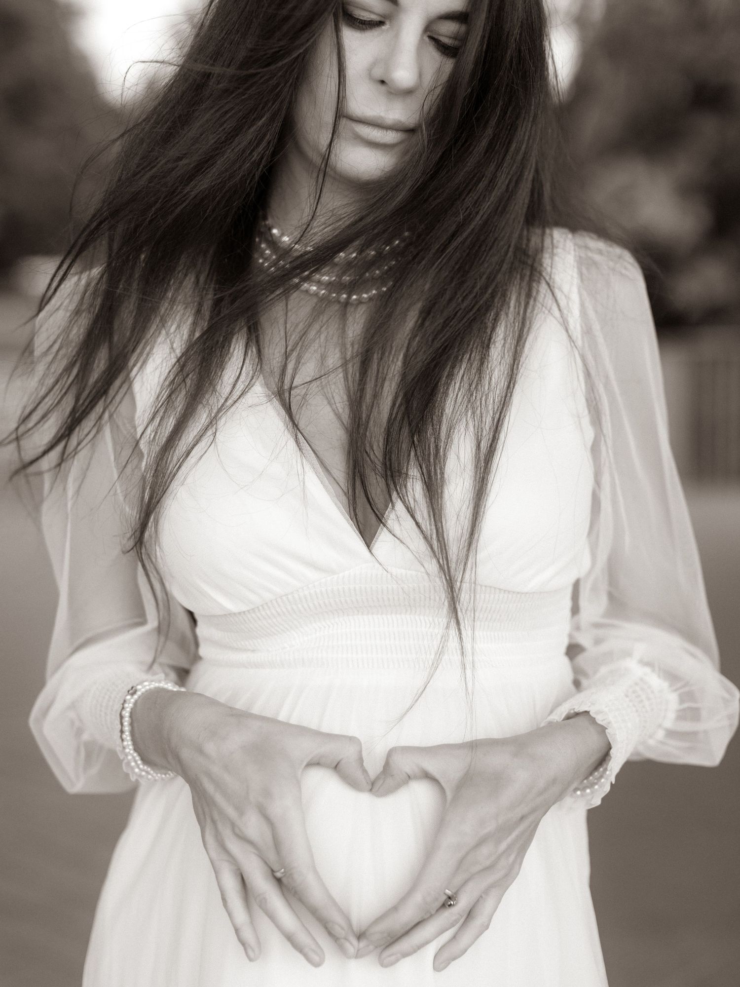 Pregnant woman in white dress making heart shape with hands on baby bump, black and white maternity photo.