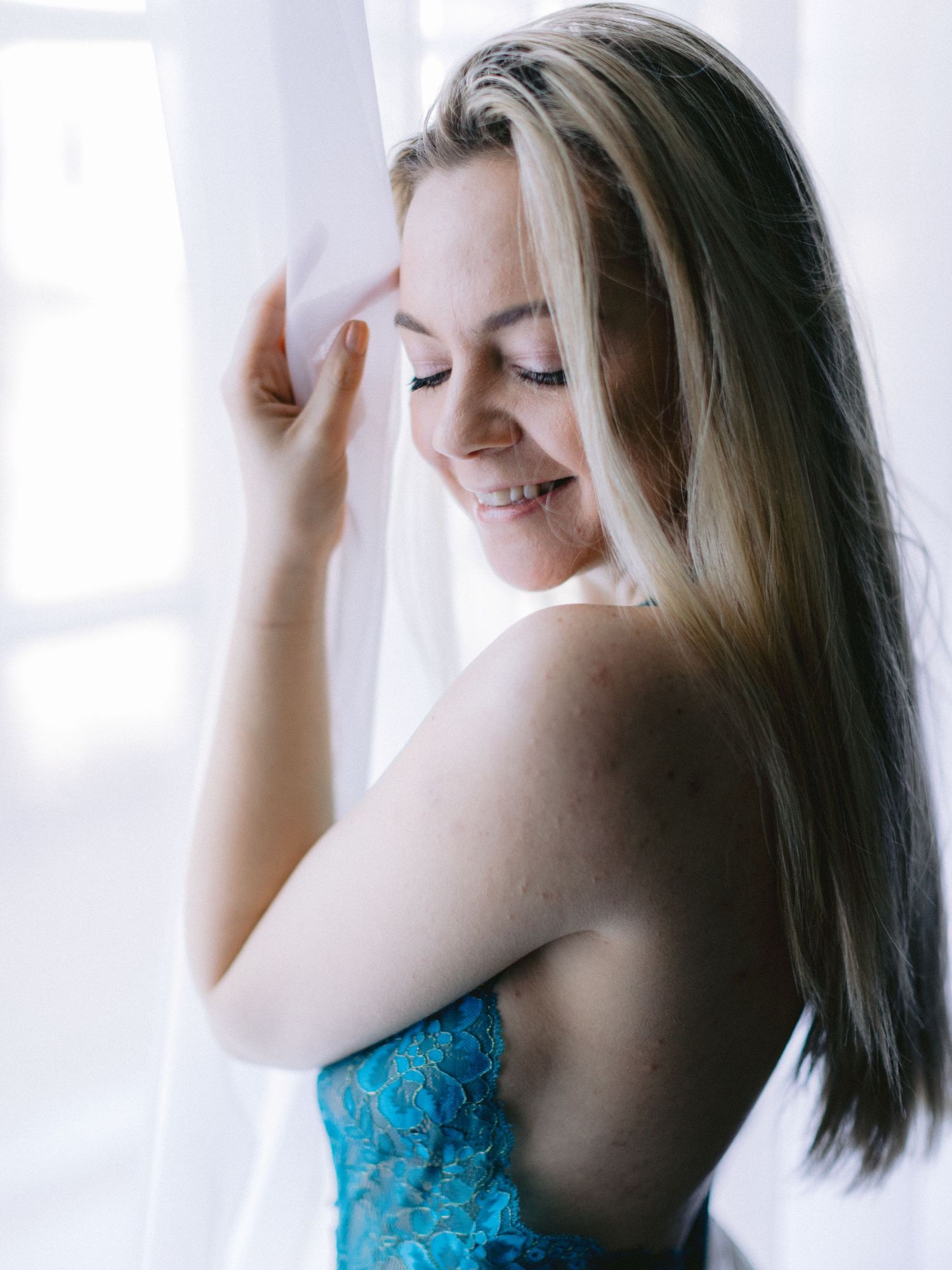Smiling blonde woman in blue lace lingerie holding a sheer white curtain near a window.