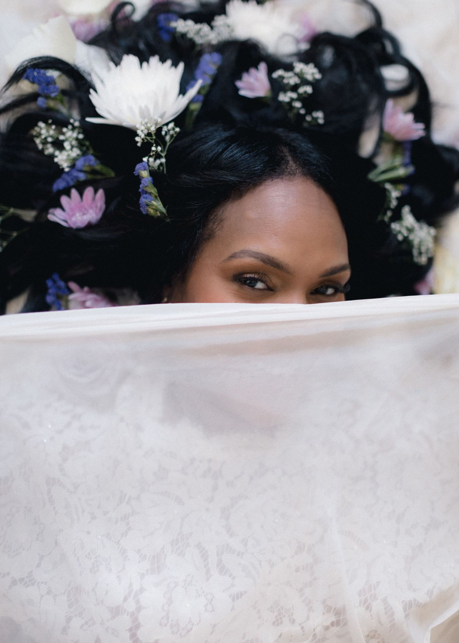 Philadelphia maternity photo session of a woman with dark hair adorned with white, purple, and blue flowers peers over a delicate veil.