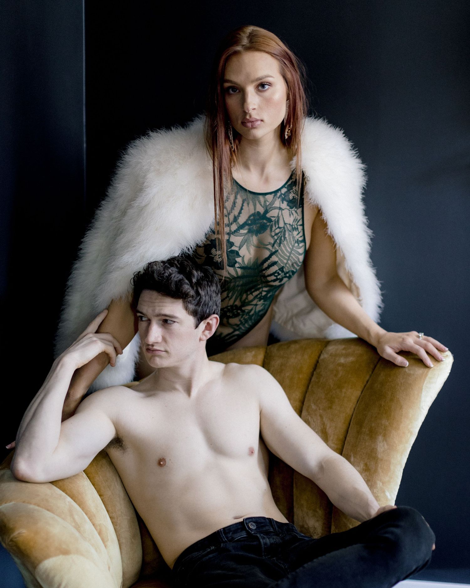 Artistic fashion portrait in dark setting with model in black lace top and white fur wrap lounging on golden chair.