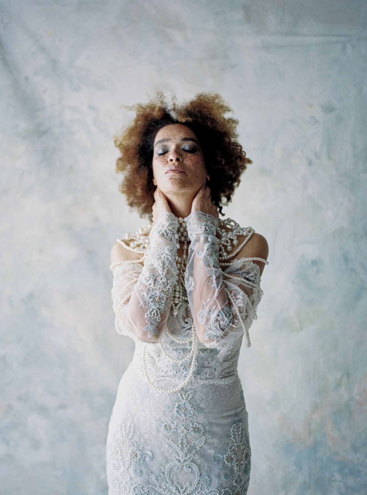 A woman in a lace bridal gown with pearl jewelry poses gracefully against a soft blue-gray backdrop.