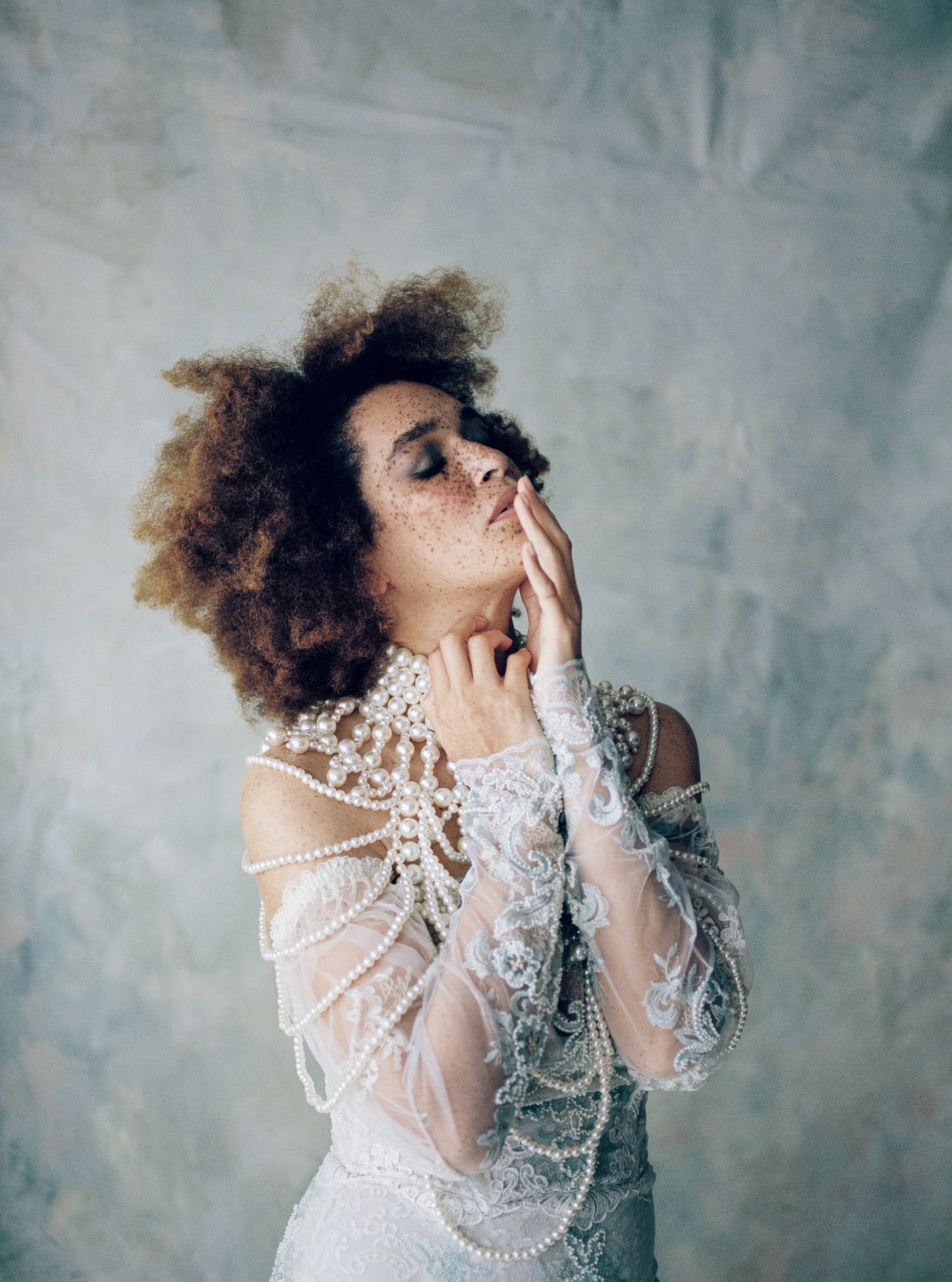 Woman in pearl-adorned white lace gown poses with hands to face against blue backdrop.
