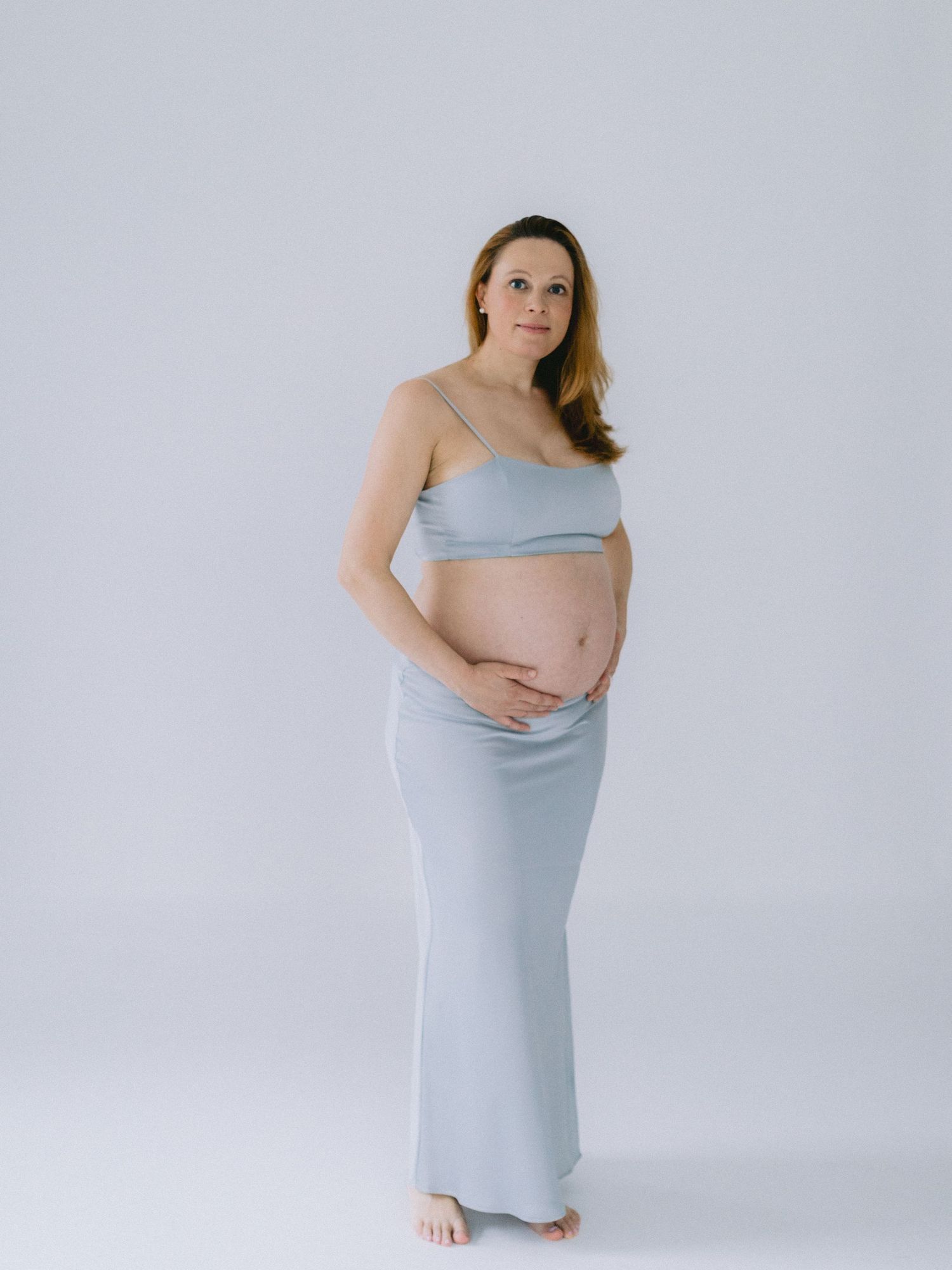 Pregnant woman in light blue crop top and maxi skirt cradling baby bump against white background.
