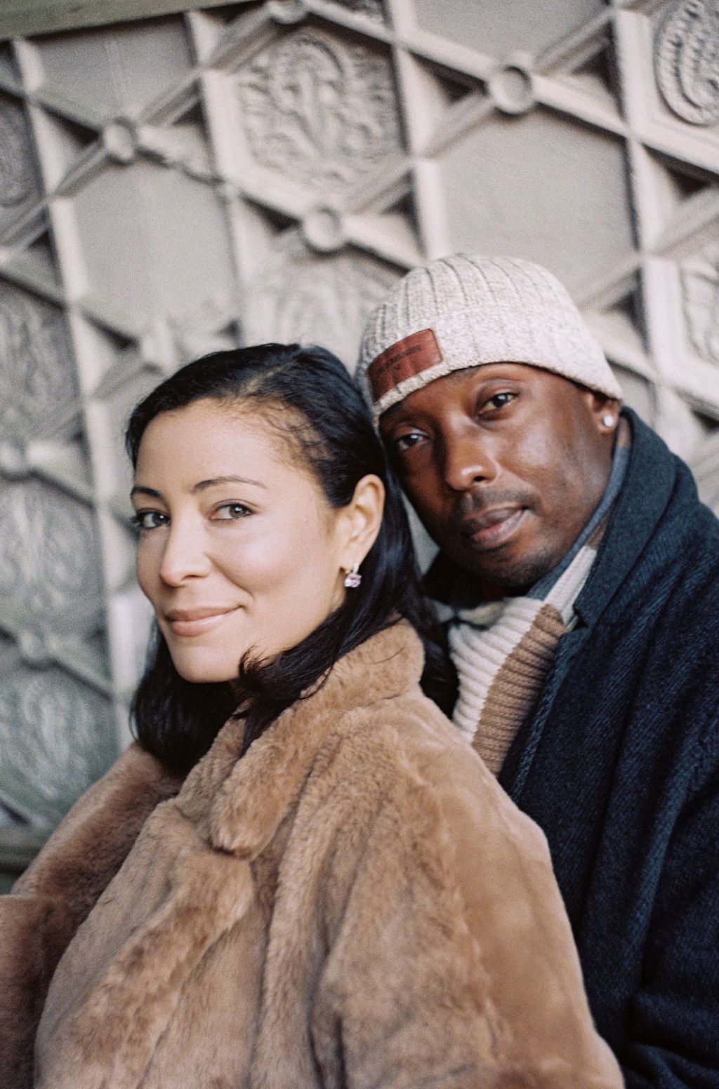 Stylish couple posing against ornate wall, woman in fur coat and man in beanie hat smiling warmly.