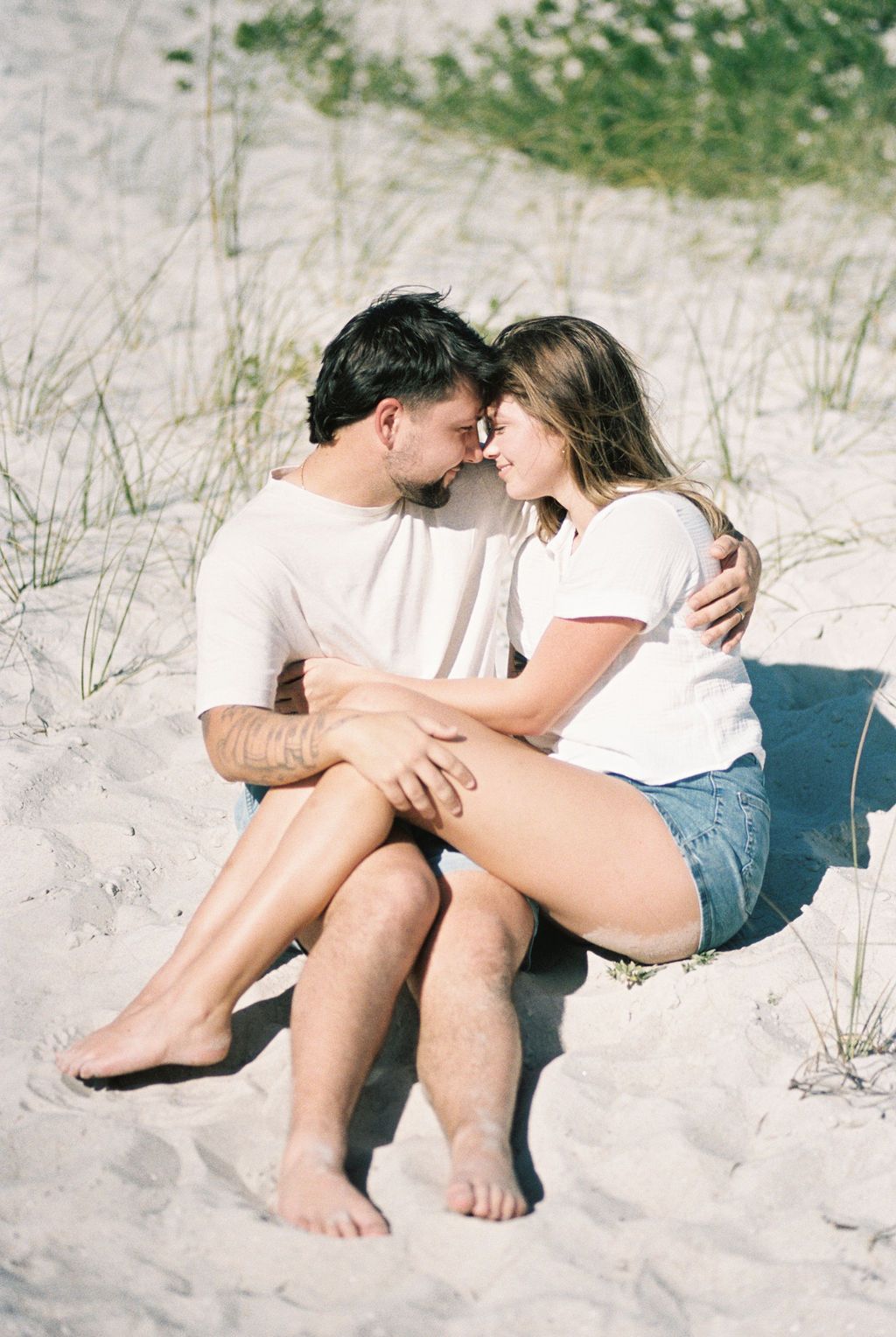 Couple in matching white tees sharing tender moment sitting together on sandy beach dunes.