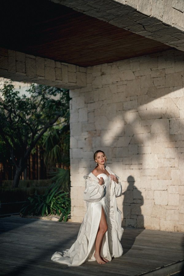 Bride in flowing white robe with thigh slit poses in sunlit stone architectural outdoor space.