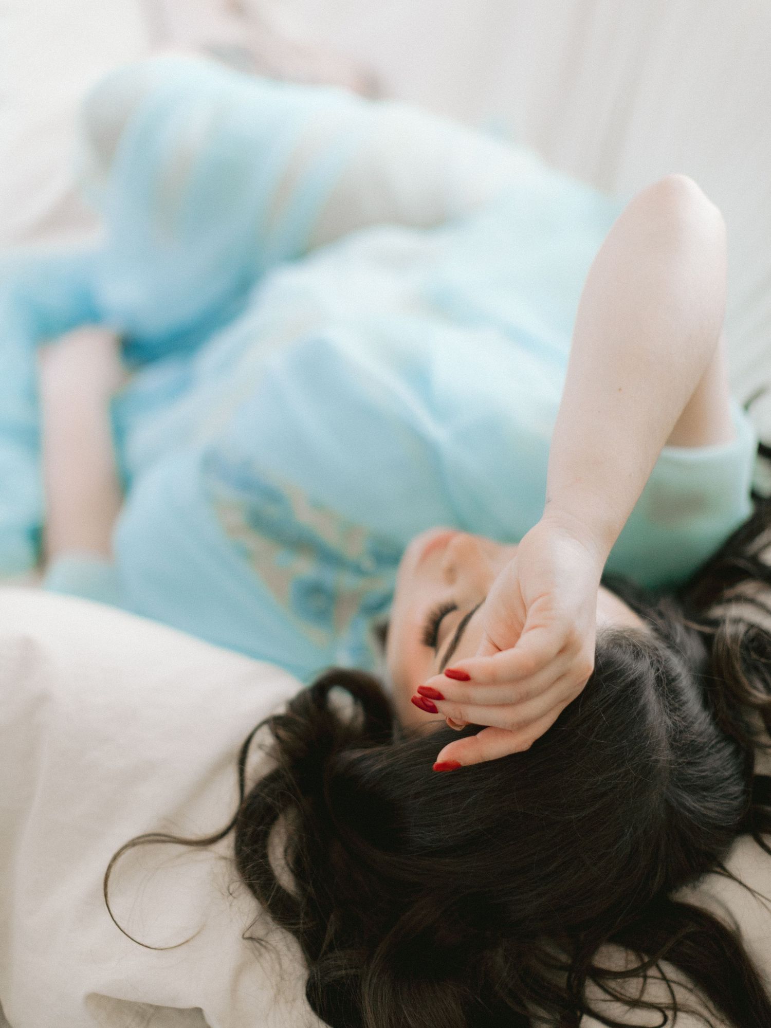 Woman in light blue dress lying down with dark hair spread out, red nails, eyes closed in dreamy pose.