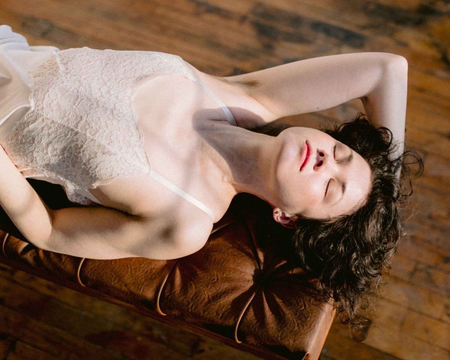 Woman in white lace lingerie lying back on a leather bench with eyes closed.