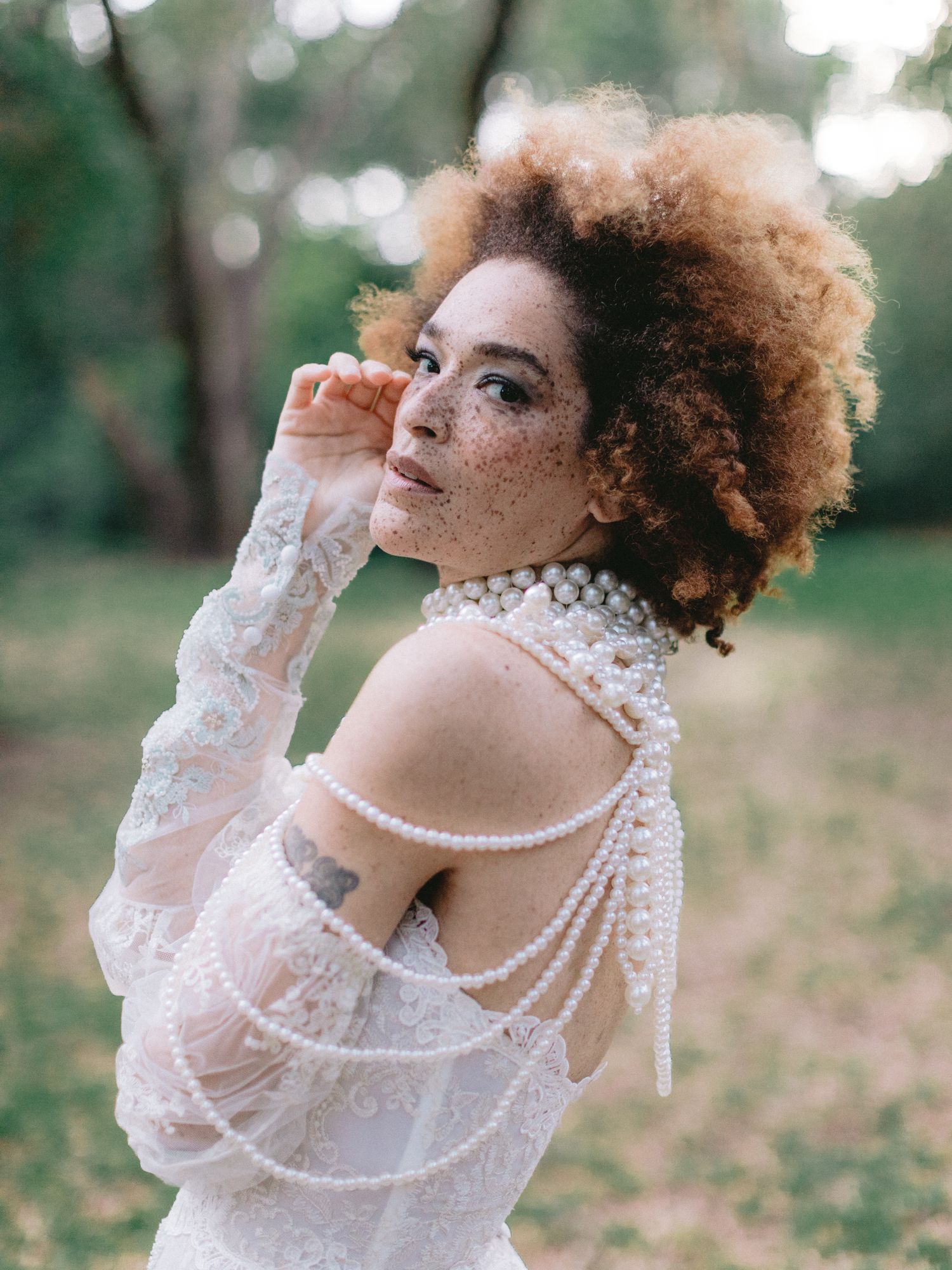 Woman with natural afro and freckles wearing pearl-draped lace bridal gown outdoors.