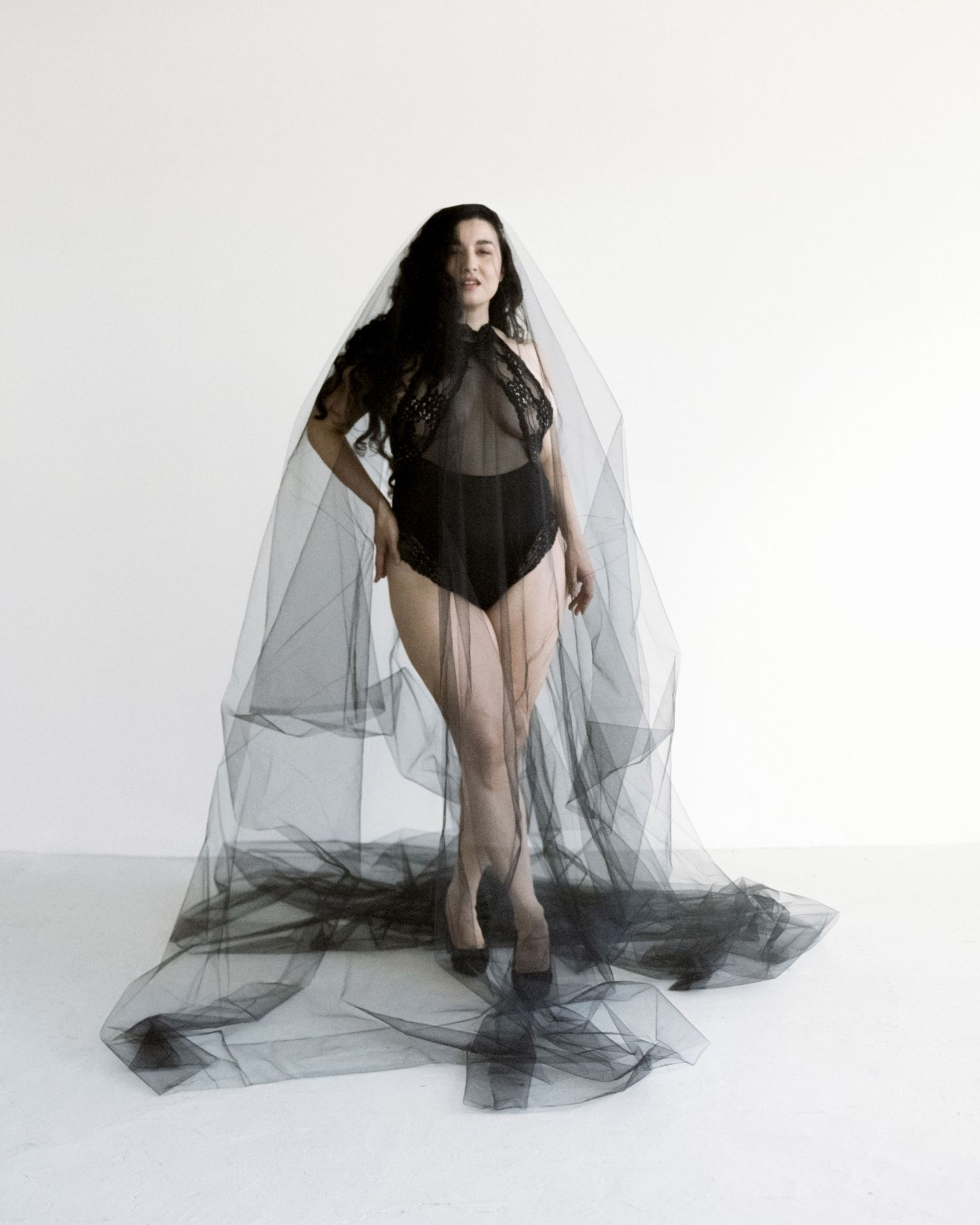 Model wearing black lace bodysuit with dramatic flowing black tulle cape in white studio.