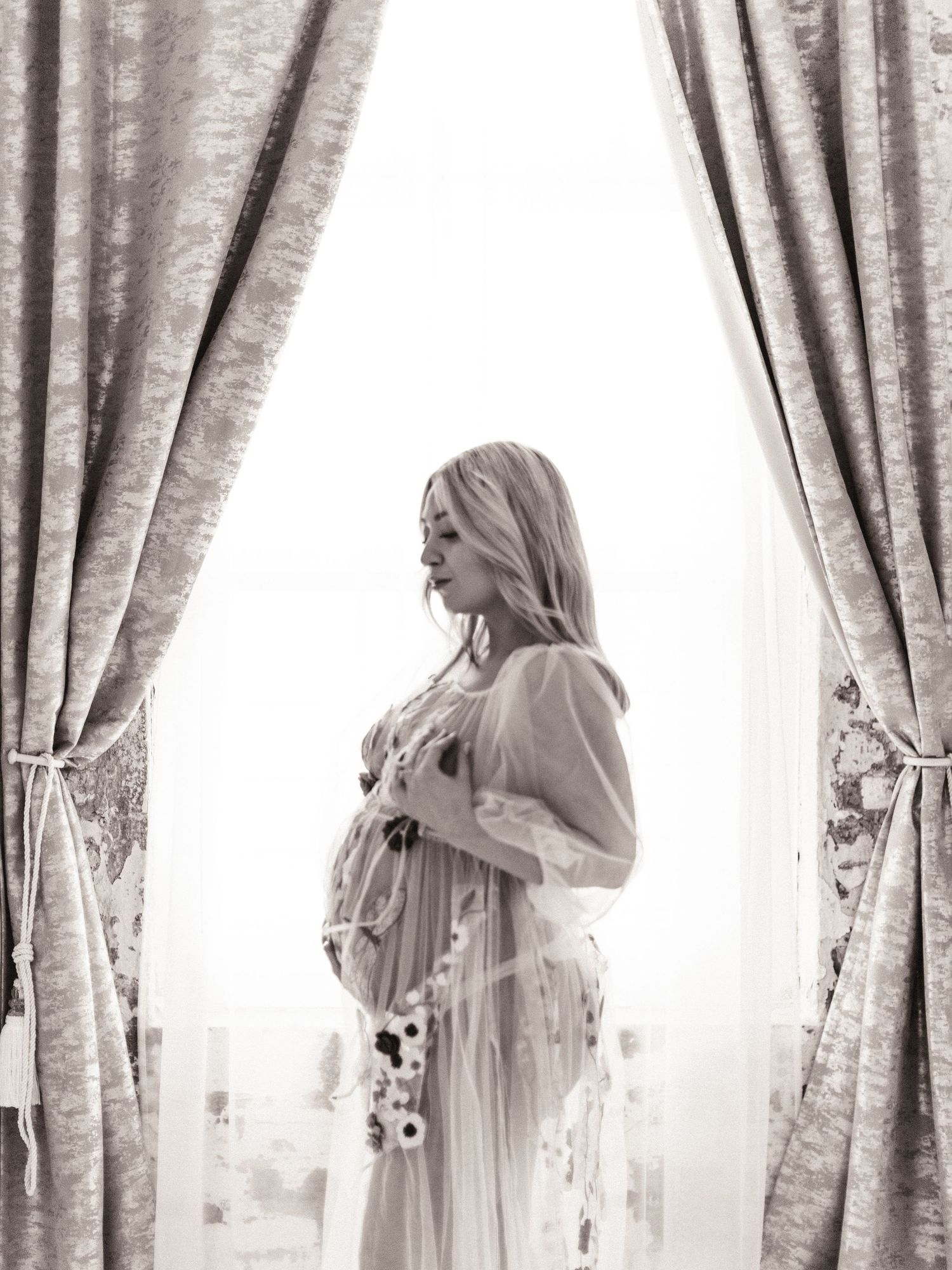 Black and white maternity photo of pregnant woman in sheer floral gown standing by elegant curtained window.