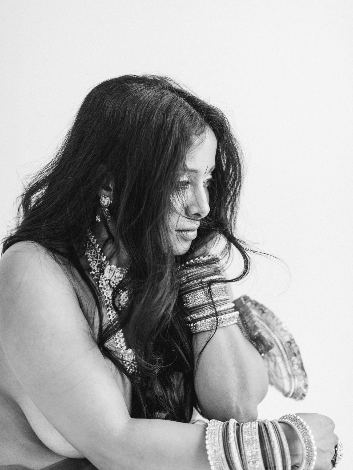 Black and white portrait of a woman with long dark hair wearing bangles and looking down in a contemplative pose.