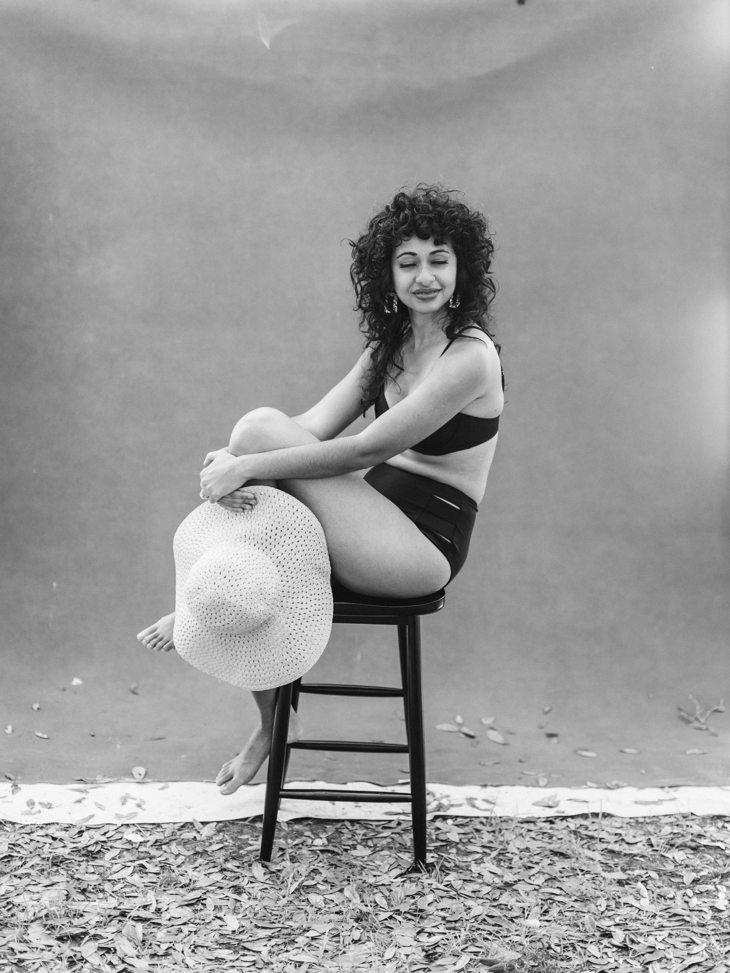 Black and white photo of a curly-haired woman in a bikini sitting on a stool with a hat.