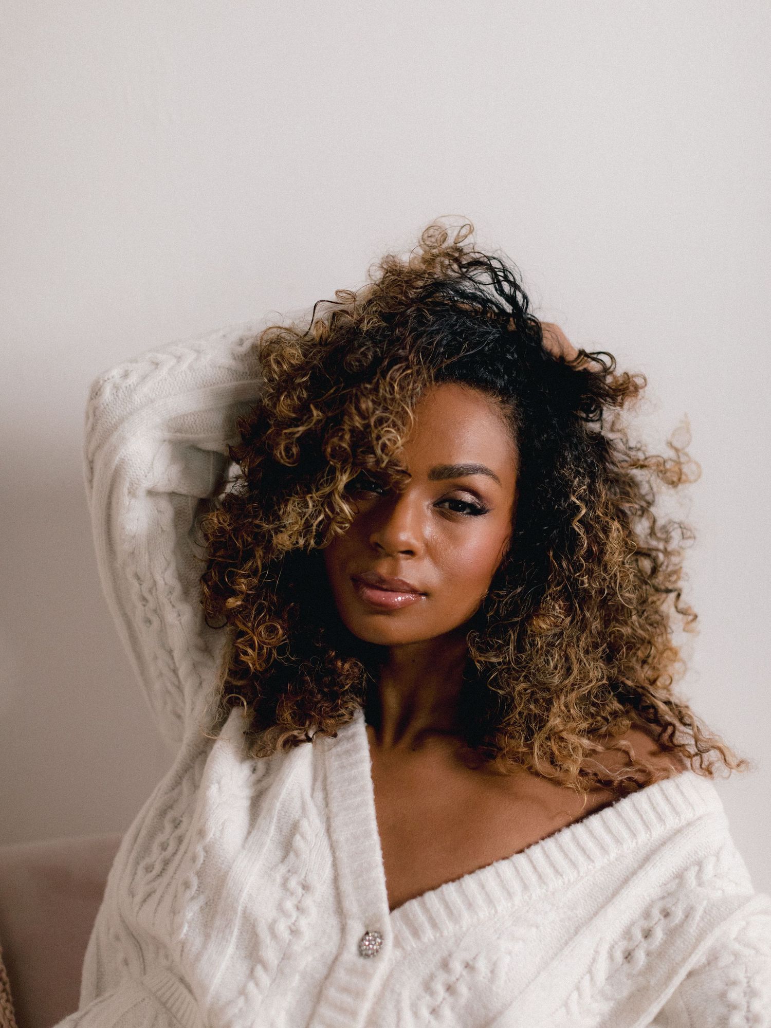 Woman with curly ombre hair wearing a white cable-knit sweater, posing against a light background.